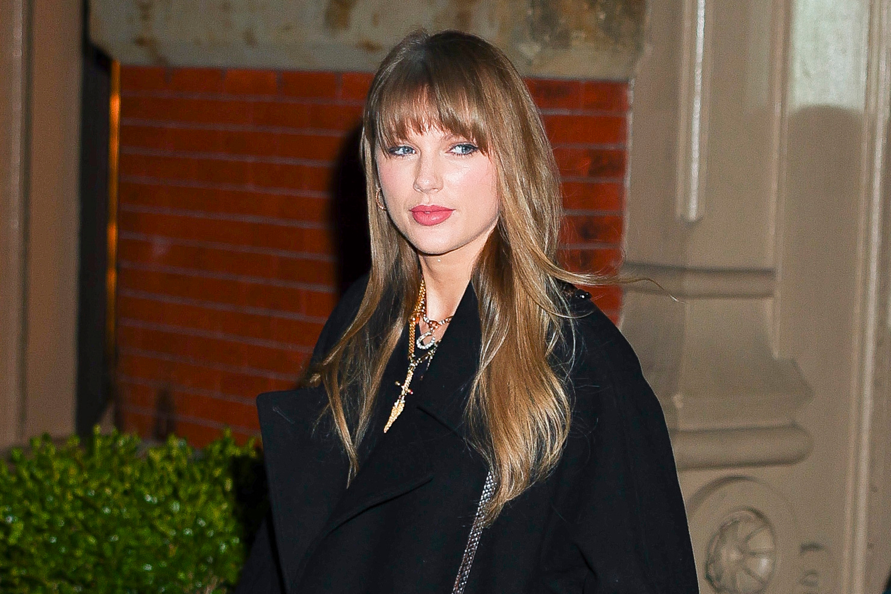 Kjendis i stilig svart kåpe med halskjede, stående utendørs. Bakgrunn av murvegg