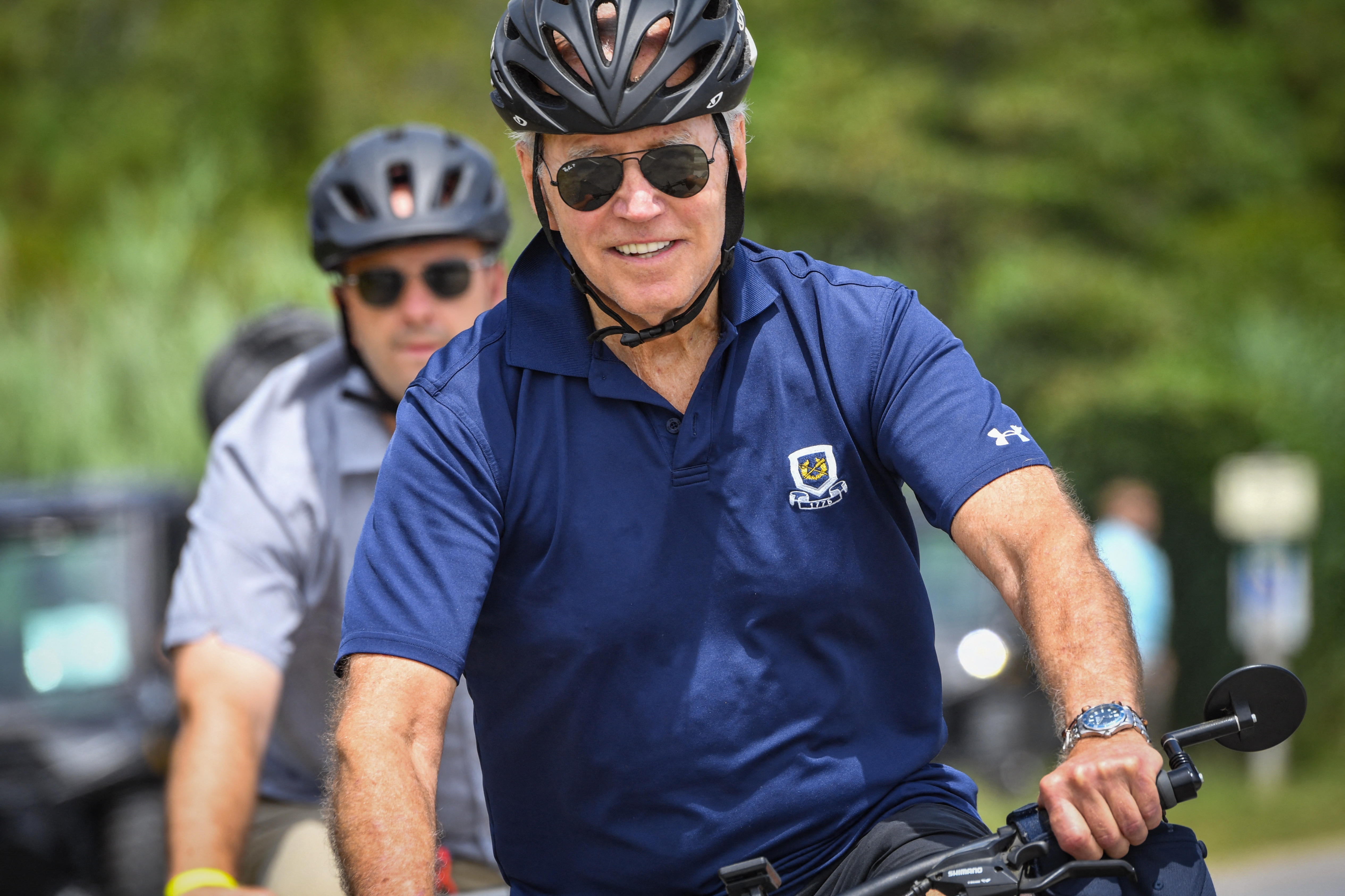 Two radical   wearing helmets thrust   bicycles outdoors. The idiosyncratic   successful  beforehand   wears a polo garment  and sunglasses, smiling portion    cycling connected  a sunny day