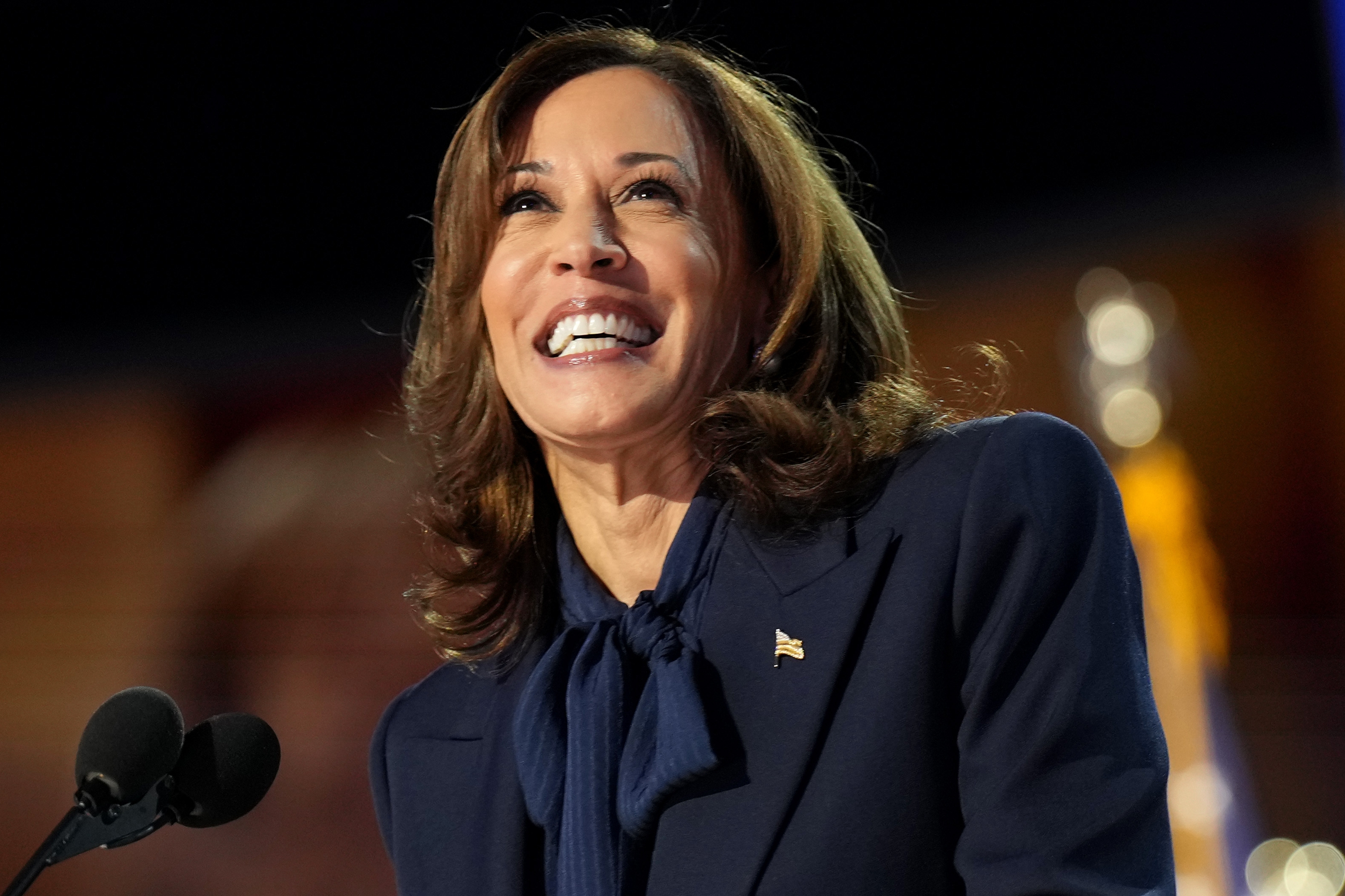 A pistillate   successful  a suit   smiles portion    speaking astatine  a podium with microphones. An American emblem  is blurred successful  the background
