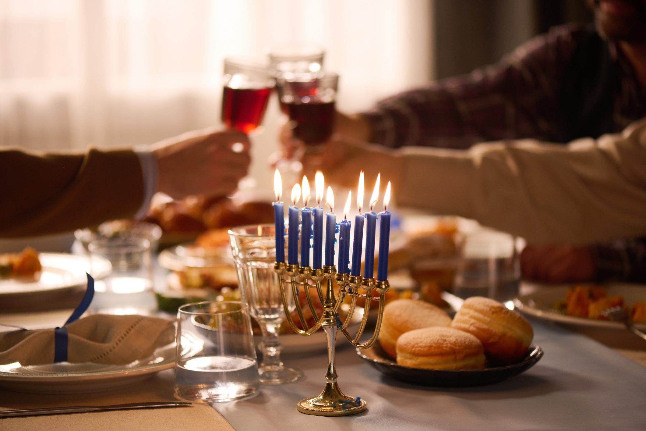 Group toasting with wine over a dining table featuring a lit menorah and a plate of traditional holiday foods