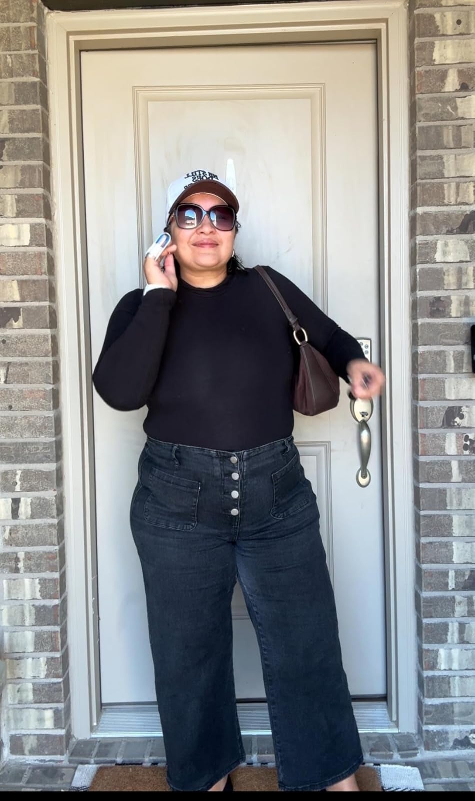 Person wearing high-waisted acheronian  bluish  jeans with a four-button waistband, achromatic  top, sunglasses, and a achromatic  shot   headdress  stands successful  beforehand   of a door, holding a telephone  and brownish  bag