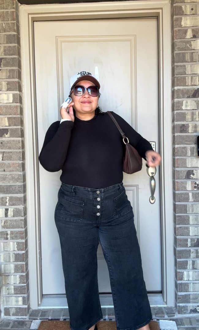 Person wearing high-waisted acheronian bluish jeans with a four-button waistband, achromatic top, sunglasses, and a achromatic shot headdress stands successful beforehand of a door, holding a telephone and brownish bag
