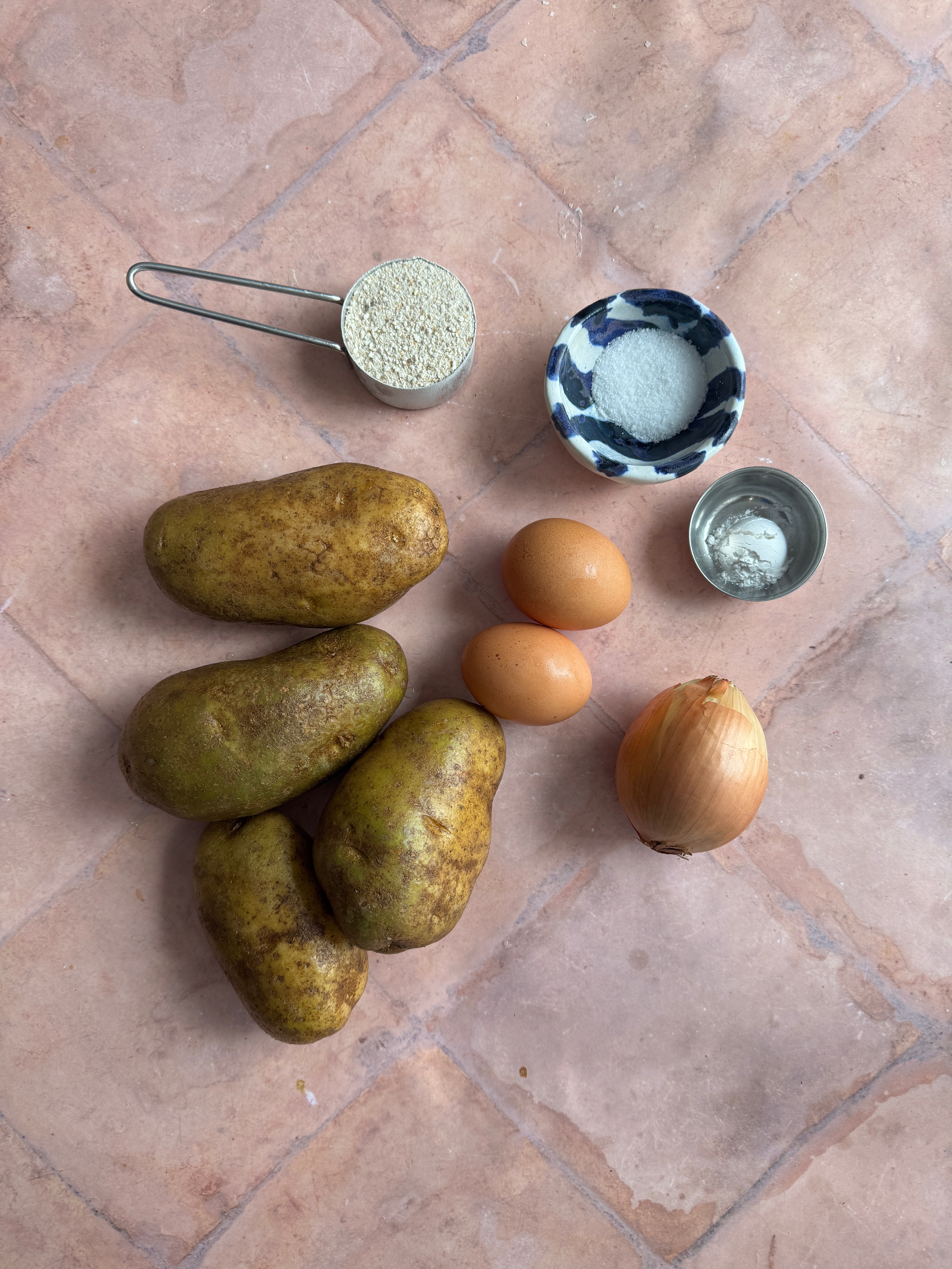 Potatoes, eggs, onion, a cup of matzo meal, a bowl of salt, and a small cup of baking powder on a tiled surface