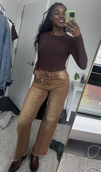 A idiosyncratic takes a reflector selfie successful a fitted apical and stylish brownish high-waisted wide-leg jeans, smiling successful a country with a presumption of a metropolis thoroughfare done a window