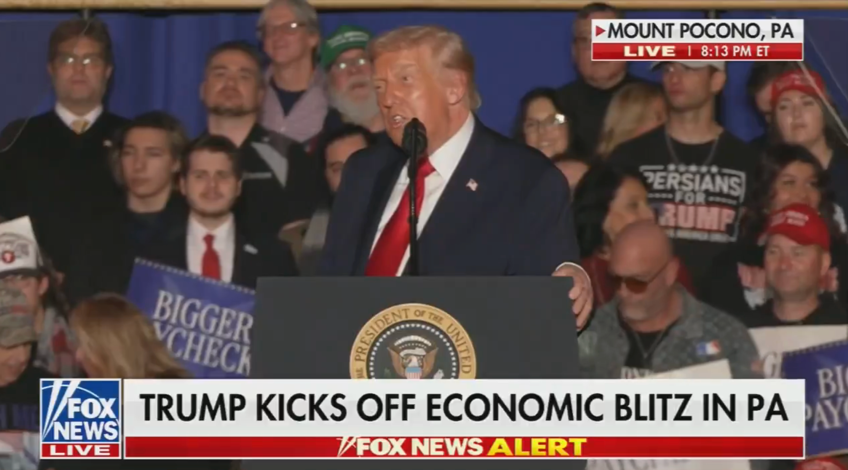 En mann taler ved et podium med en presidentsegl, omgitt av et publikum som holder skilt ved et rallyarrangement. Nyhetsticker lyder: 