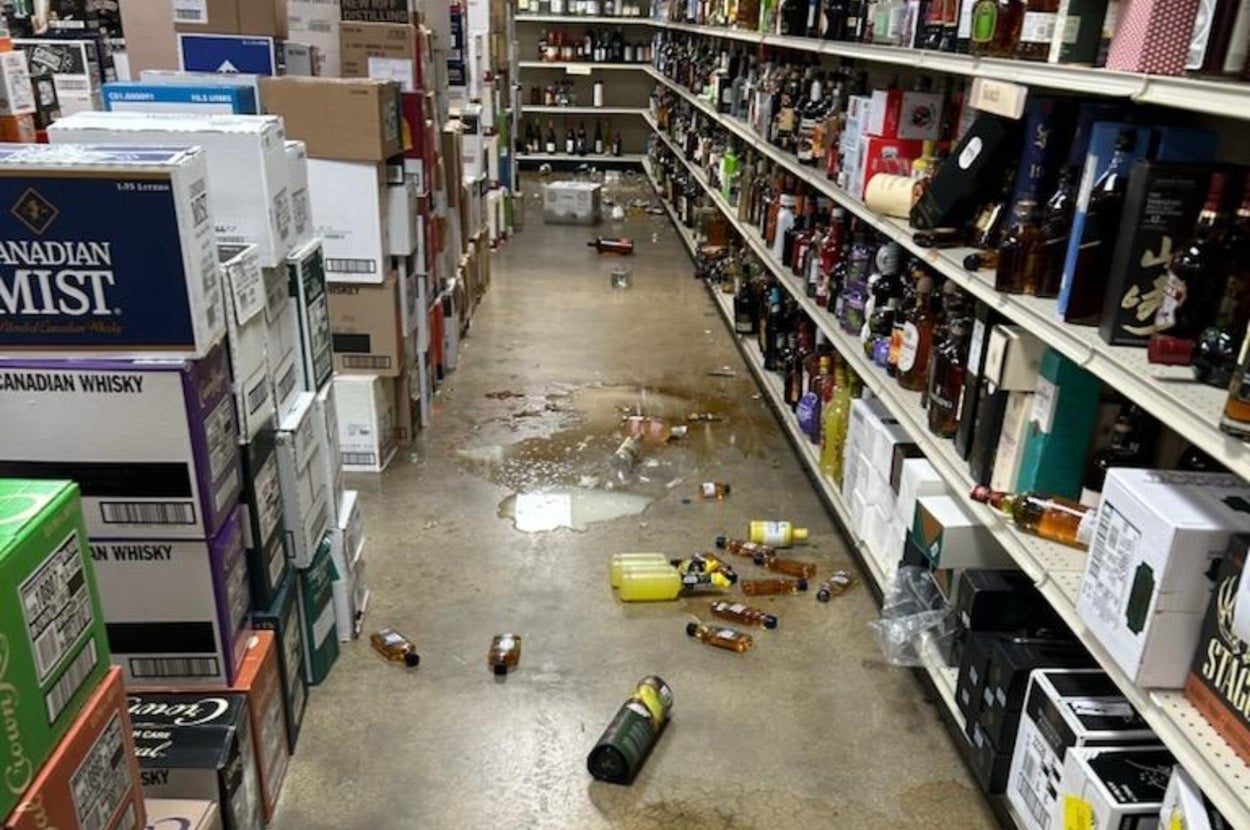 店の通路に落ちた割れたボトルと液体、棚にアルコール類が並ぶ。