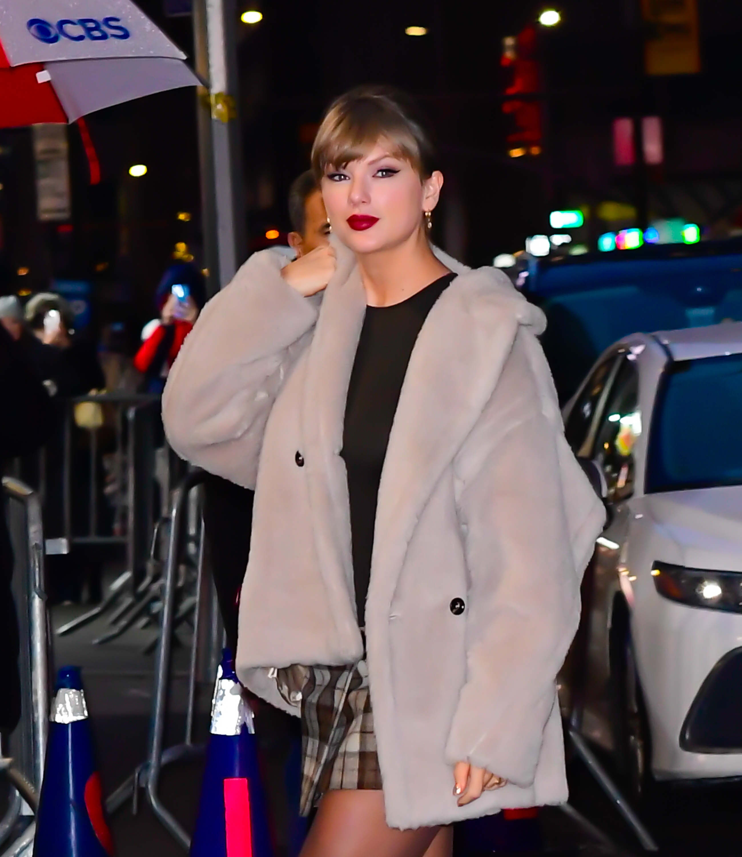 Person successful  chic fur coat, plaid mini skirt, and acheronian  apical  connected  metropolis  thoroughfare  astatine  night, walking past   vehicles and onlookers