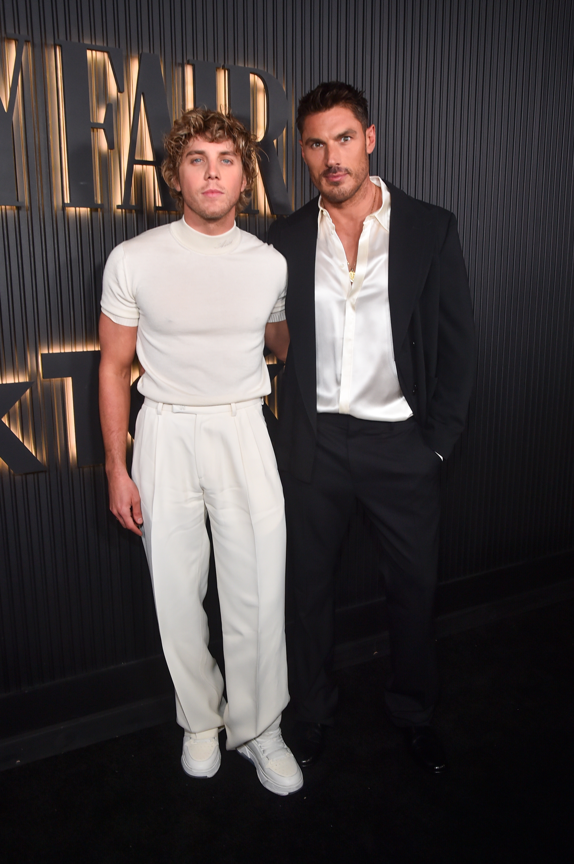 Two individuals airs  astatine  a Vanity Fair event. One wears a casual t-shirt and trousers, the different   a ceremonial  blazer with an open-neck shirt