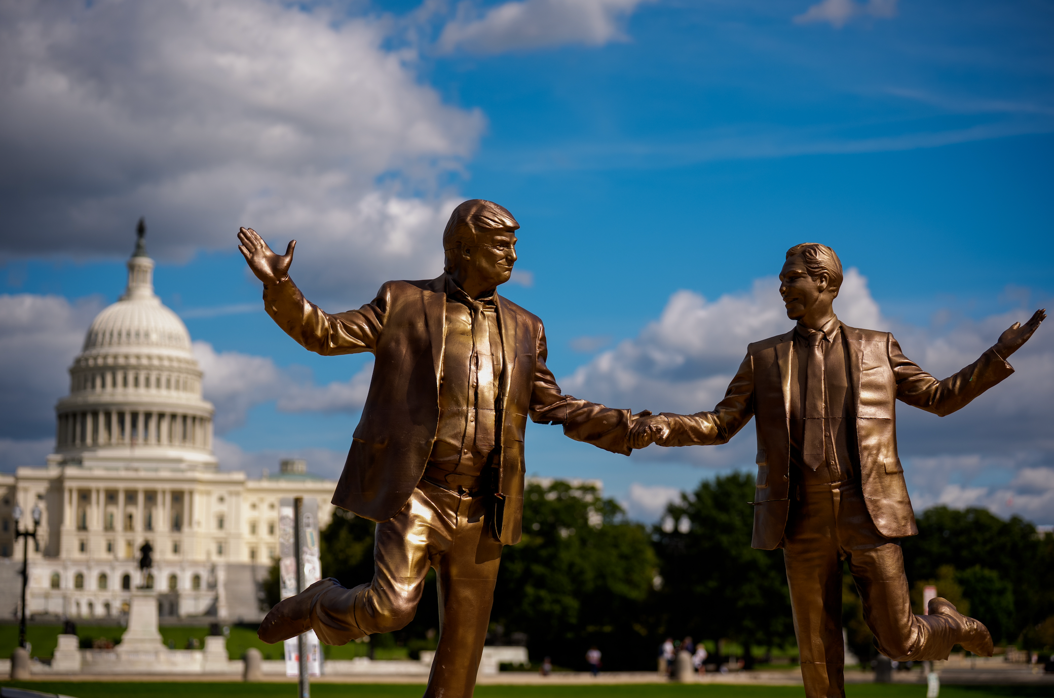 Two bronze statues of men successful suits joyfully holding hands adjacent the U.S. Capitol building