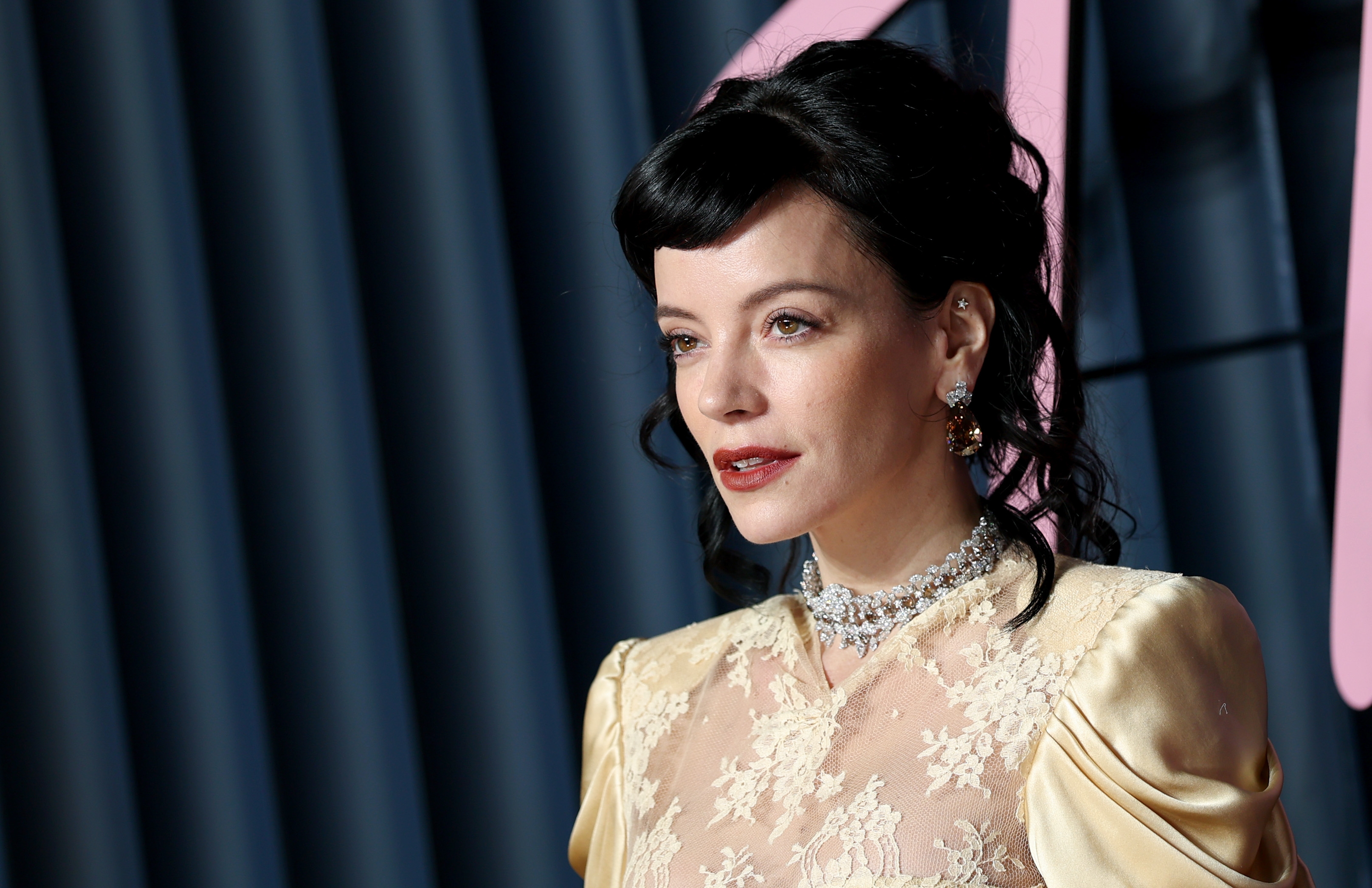 Person connected  the reddish  carpet wearing a lace gown with intricate designs, accessorized with earrings and layered necklaces