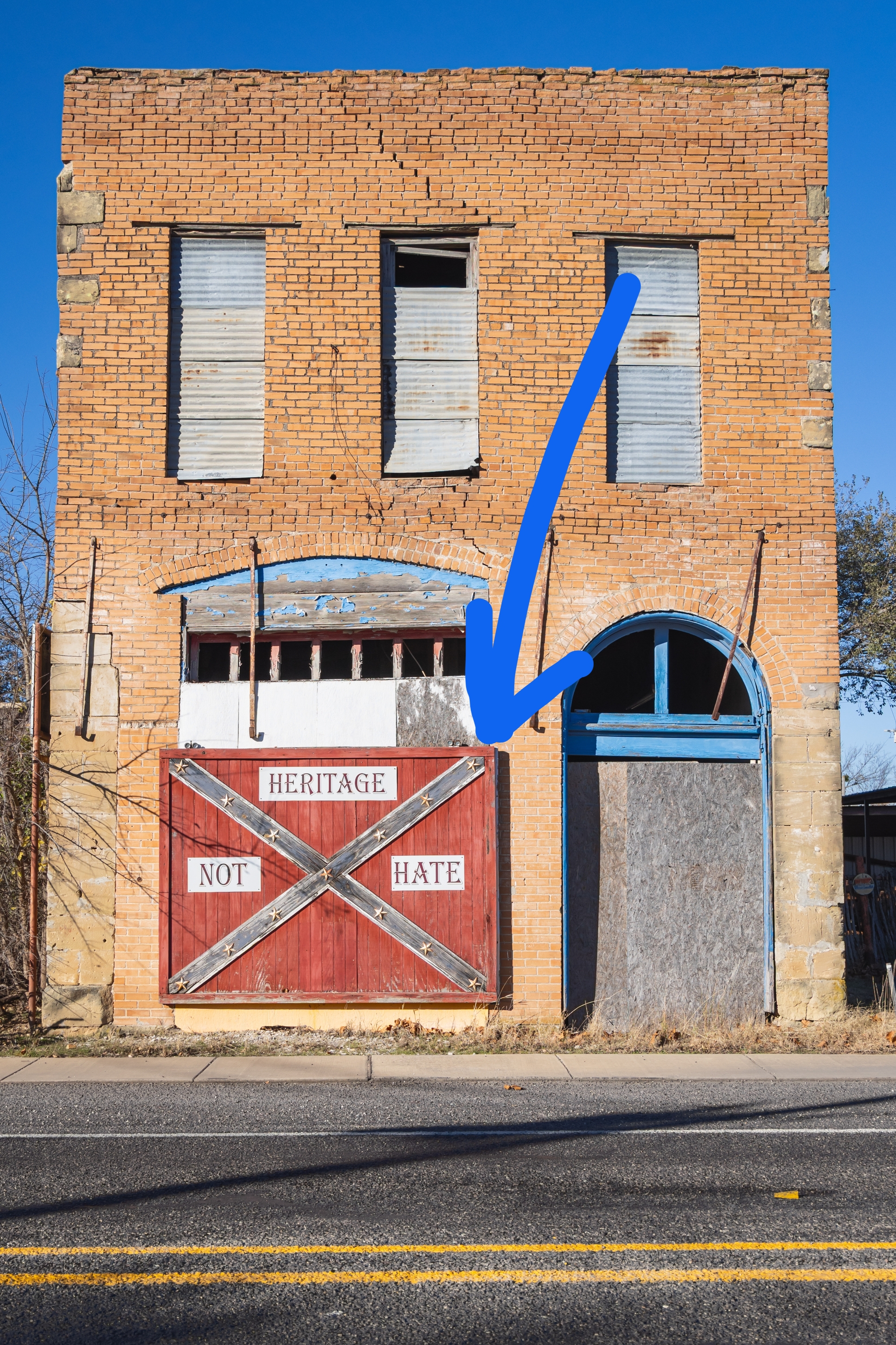 Abandoned two-story ceramic  gathering  with boarded windows and a reddish  doorway  displaying the substance   "Heritage Not Hate."