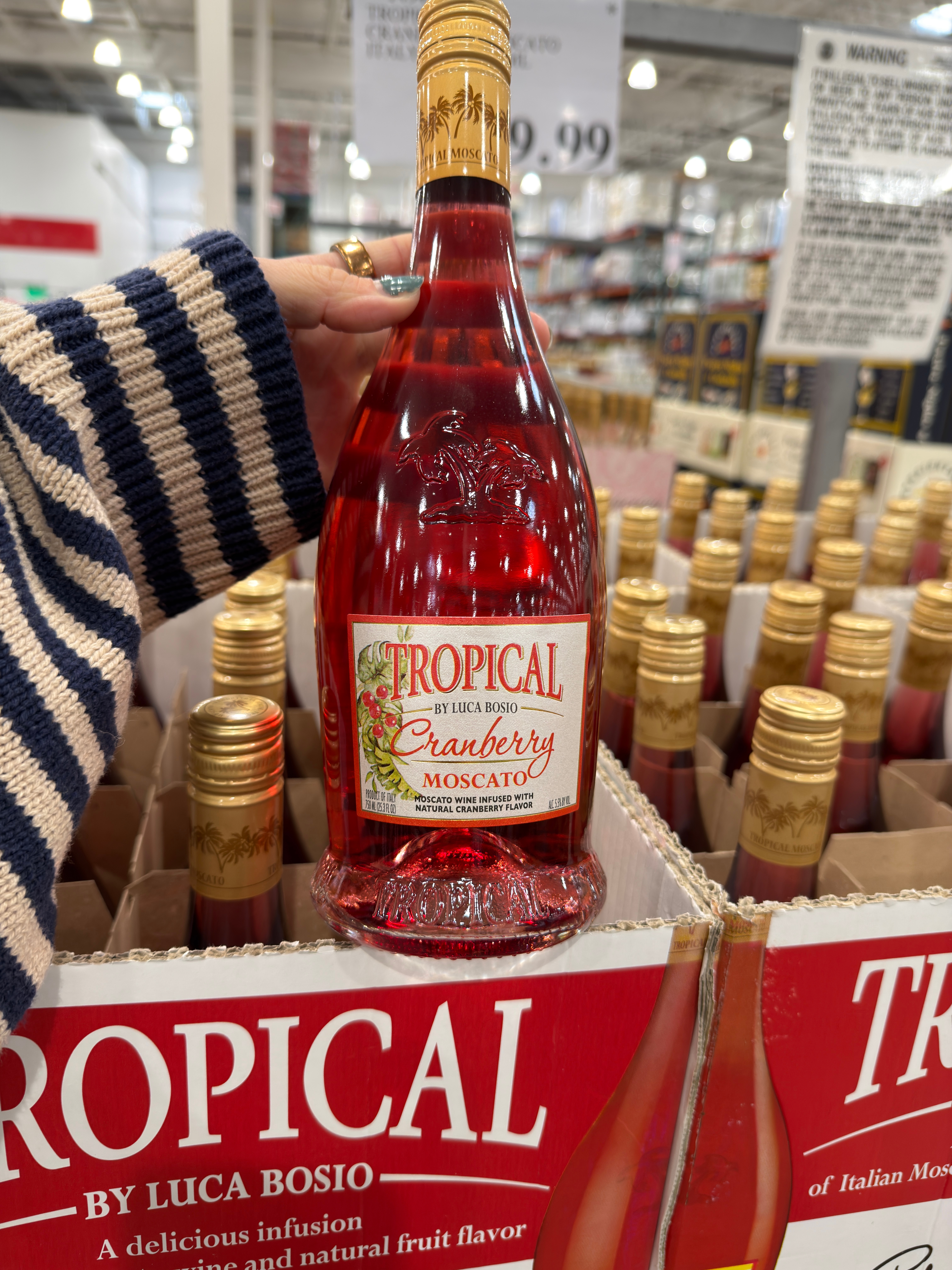 A hand holding a bottle of Tropical by Luca Bosio Cranberry Moscato wine in a store, with boxes of the same wine in the background