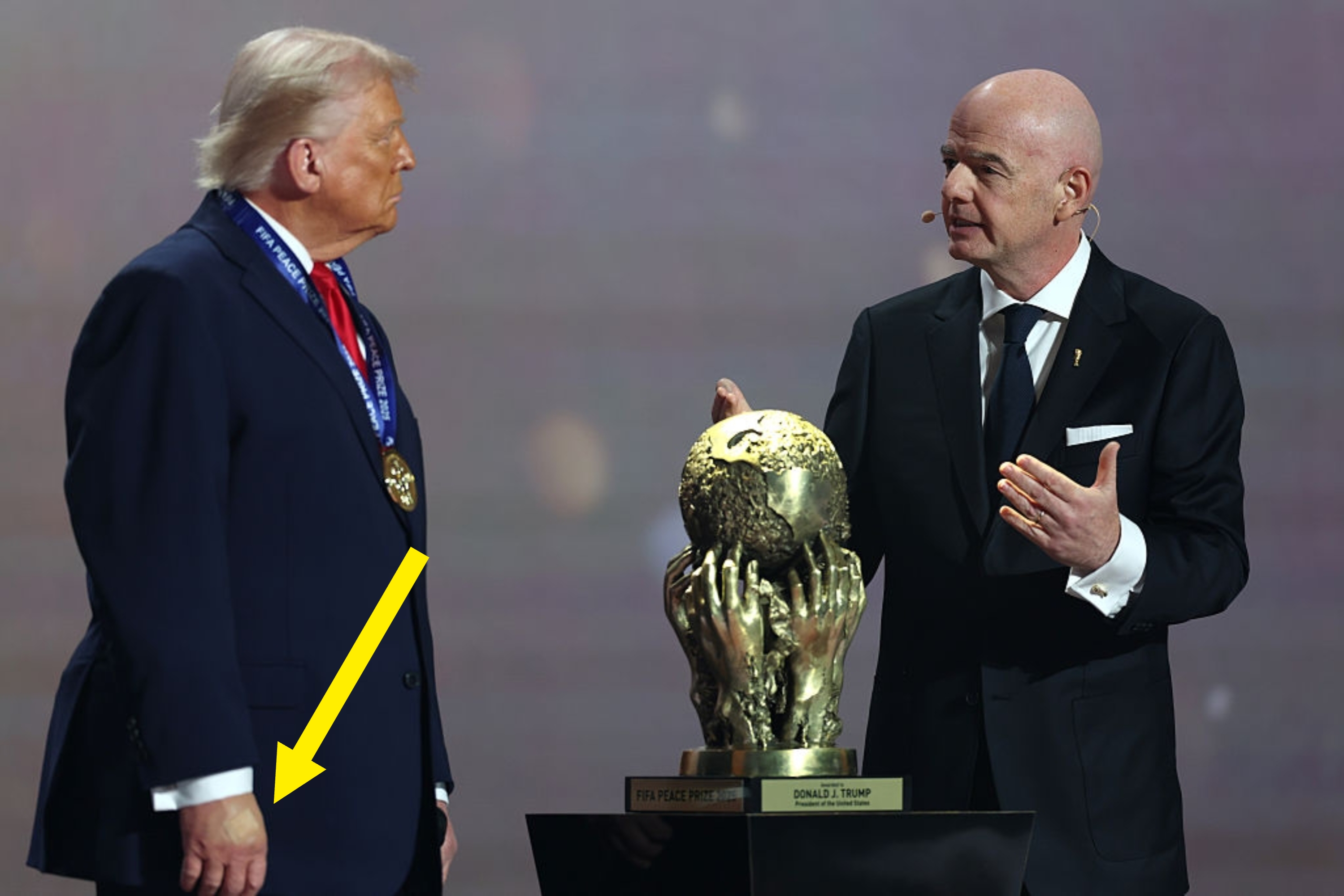 To menn i dress diskuterer et fotballpokal i gull på et podium. En mann bærer en medalje, og den andre gestikulerer mot trofeet
