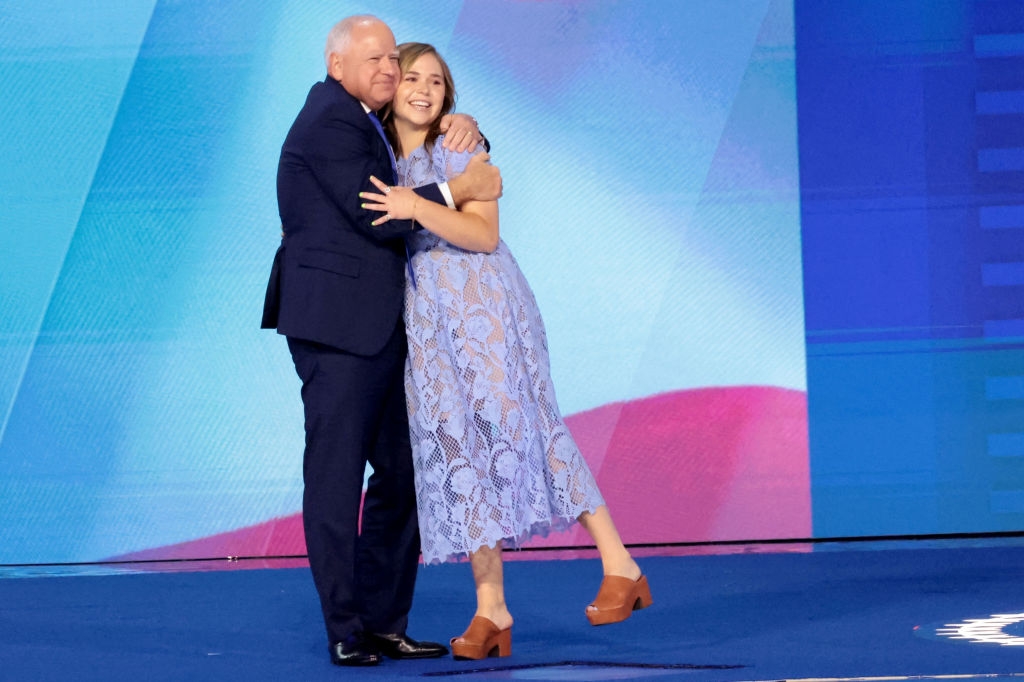 To individer omfavner på scenen ved et arrangement. Den ene har på seg dress, og den andre er i blondekjole med platåsko