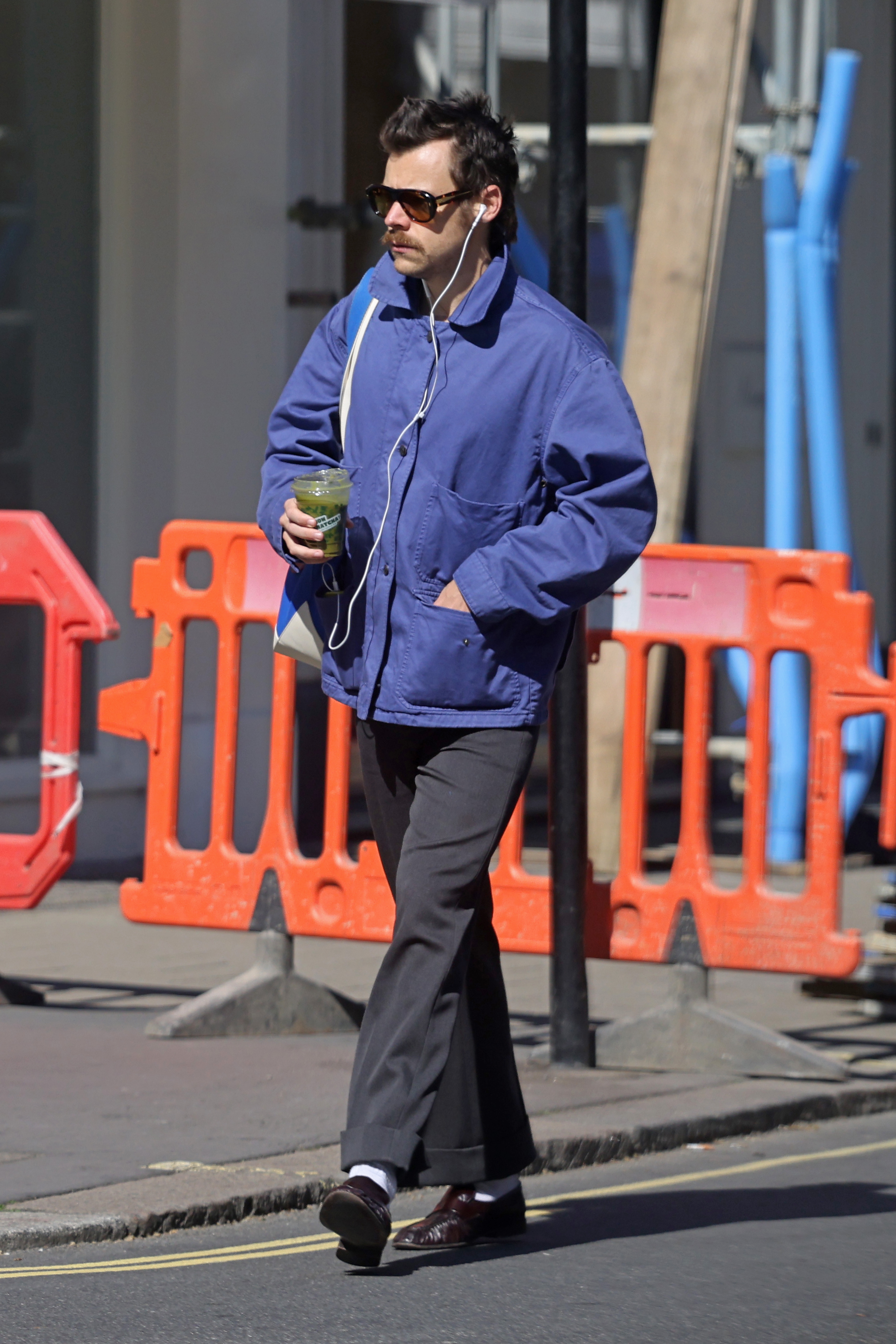 Person walking connected  a metropolis  street, wearing a casual jacket, trousers, sunglasses, and earphones, holding a drink. Construction barriers are successful  the background