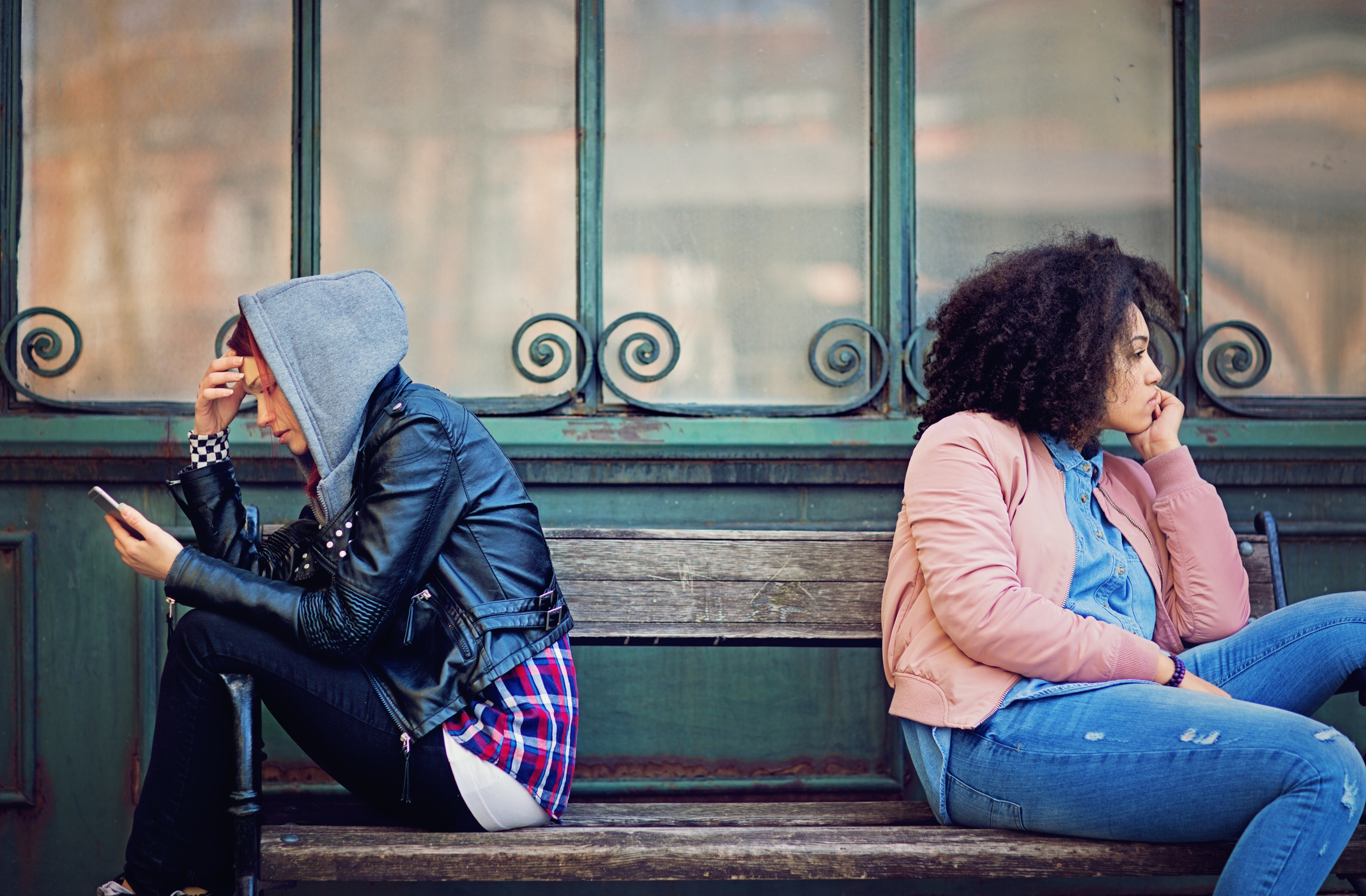 Two radical   are sitting connected  a bench, backmost  to back. One is utilizing a phone, wearing a hoodie and leather jacket. The different   looks away, appearing contemplative