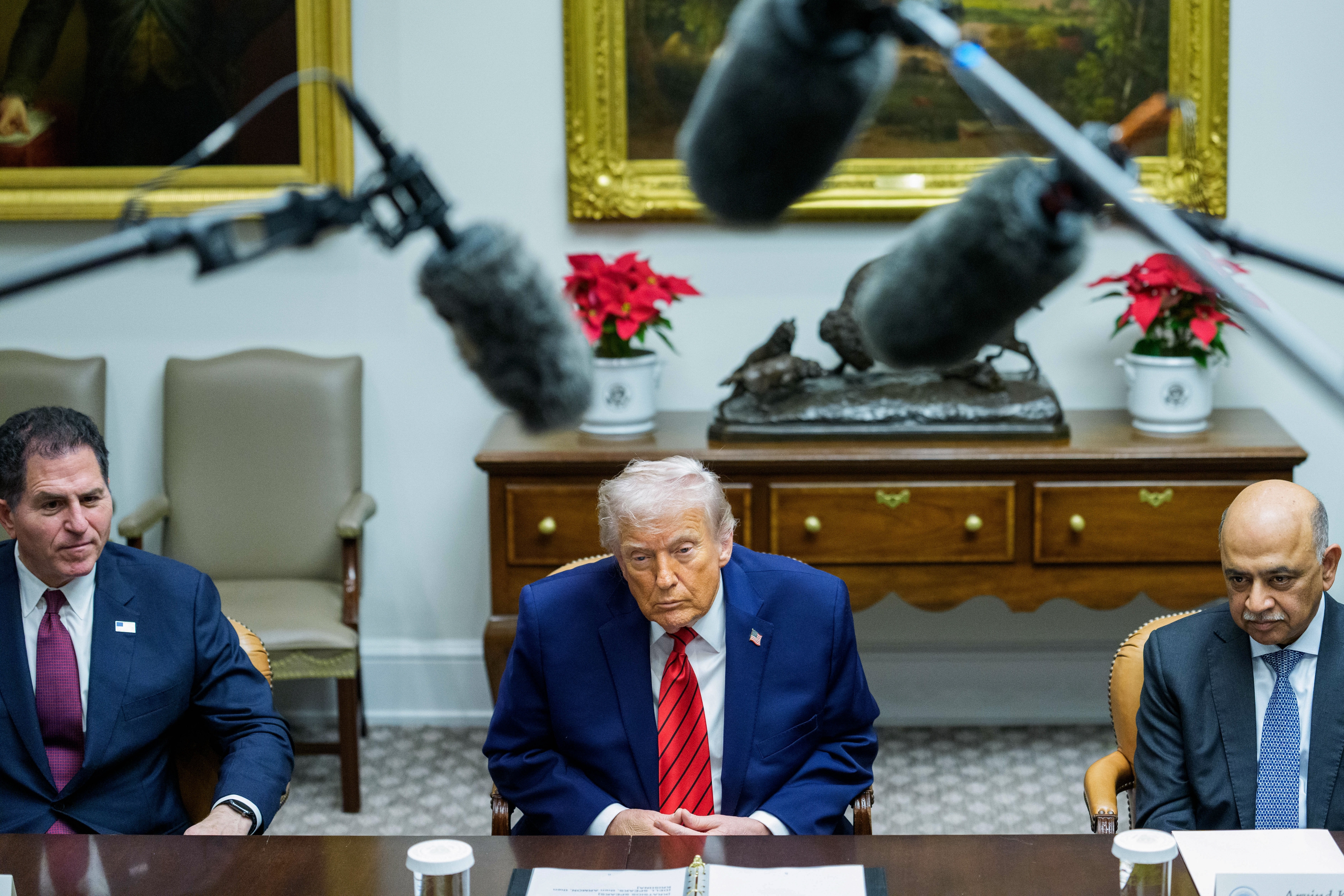 Tre menn sitter ved et konferansebord med mikrofoner over. Mannen i sentrum har på seg blå dress og rødt slips. Innstillingen er formell