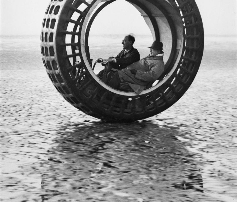 Two men sitting wrong  a large, wheel-like monowheel conveyance  connected  a creaseless  surface, facing sideways