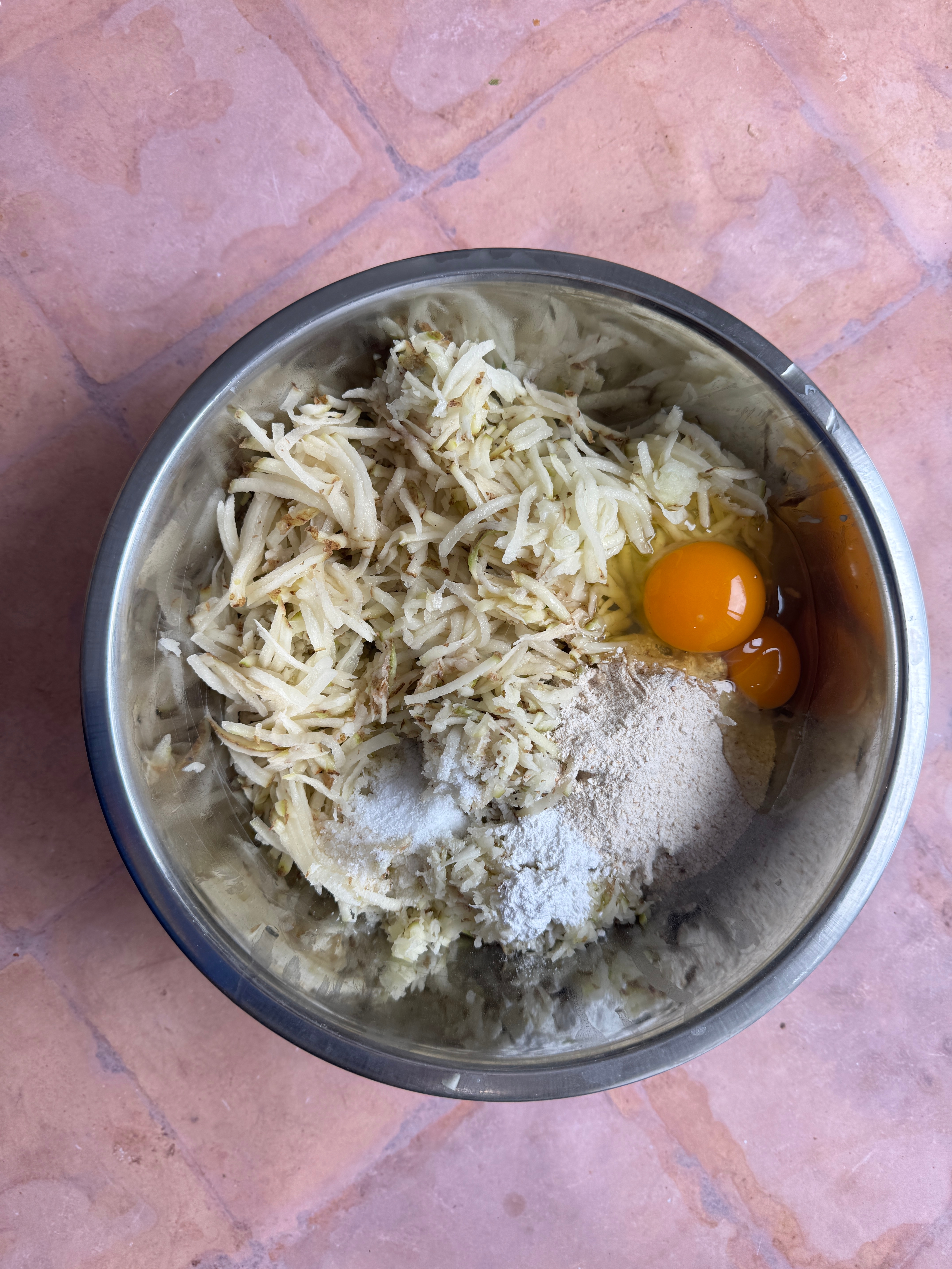 Grated potatoes, an egg, matzo meal, and seasoning in a metal bowl,  being prepared for cooking