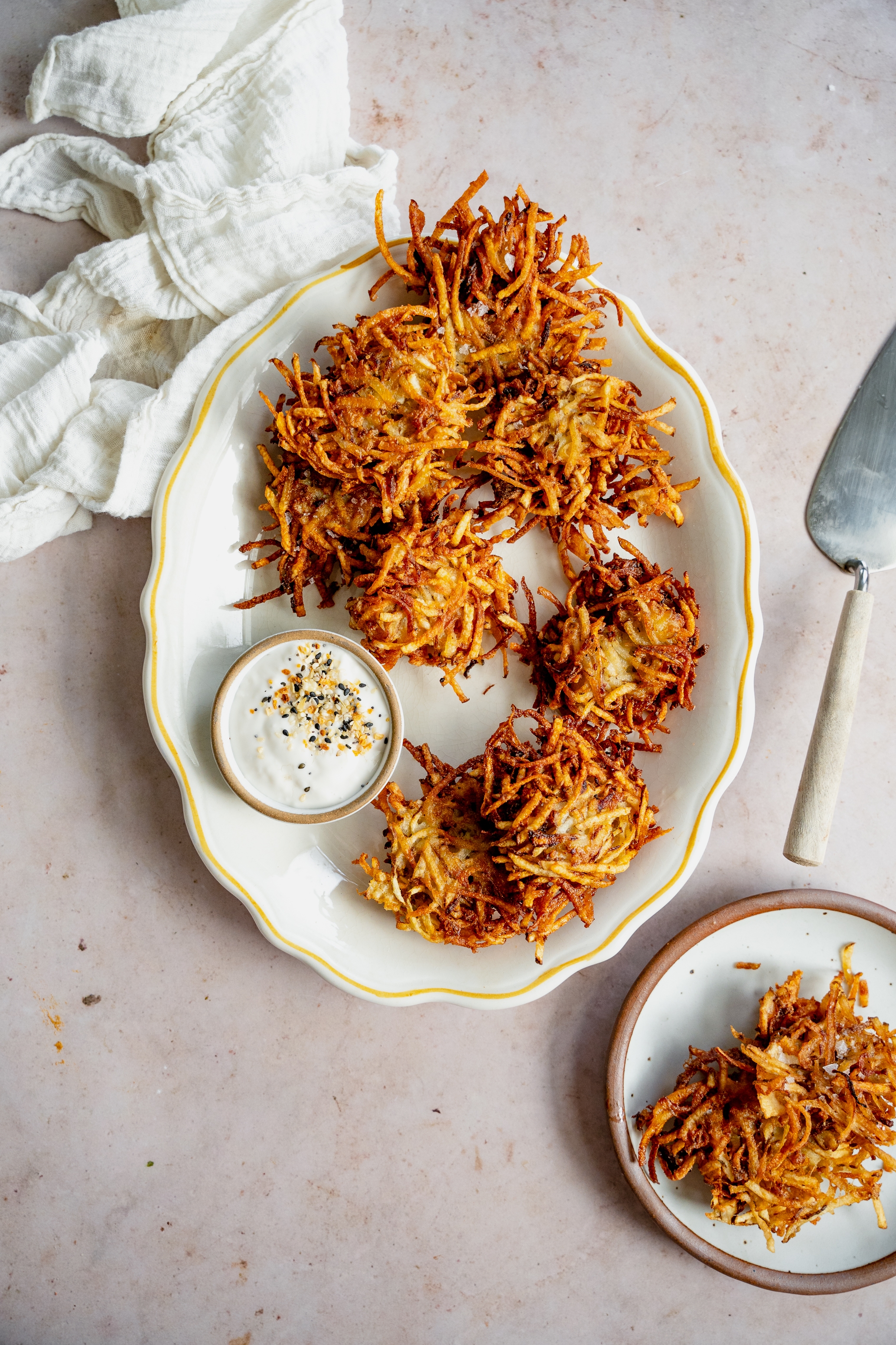 Plate of crispy hash browns with a small dish of creamy dip, served on a light surface with a white cloth and metal spatula nearby