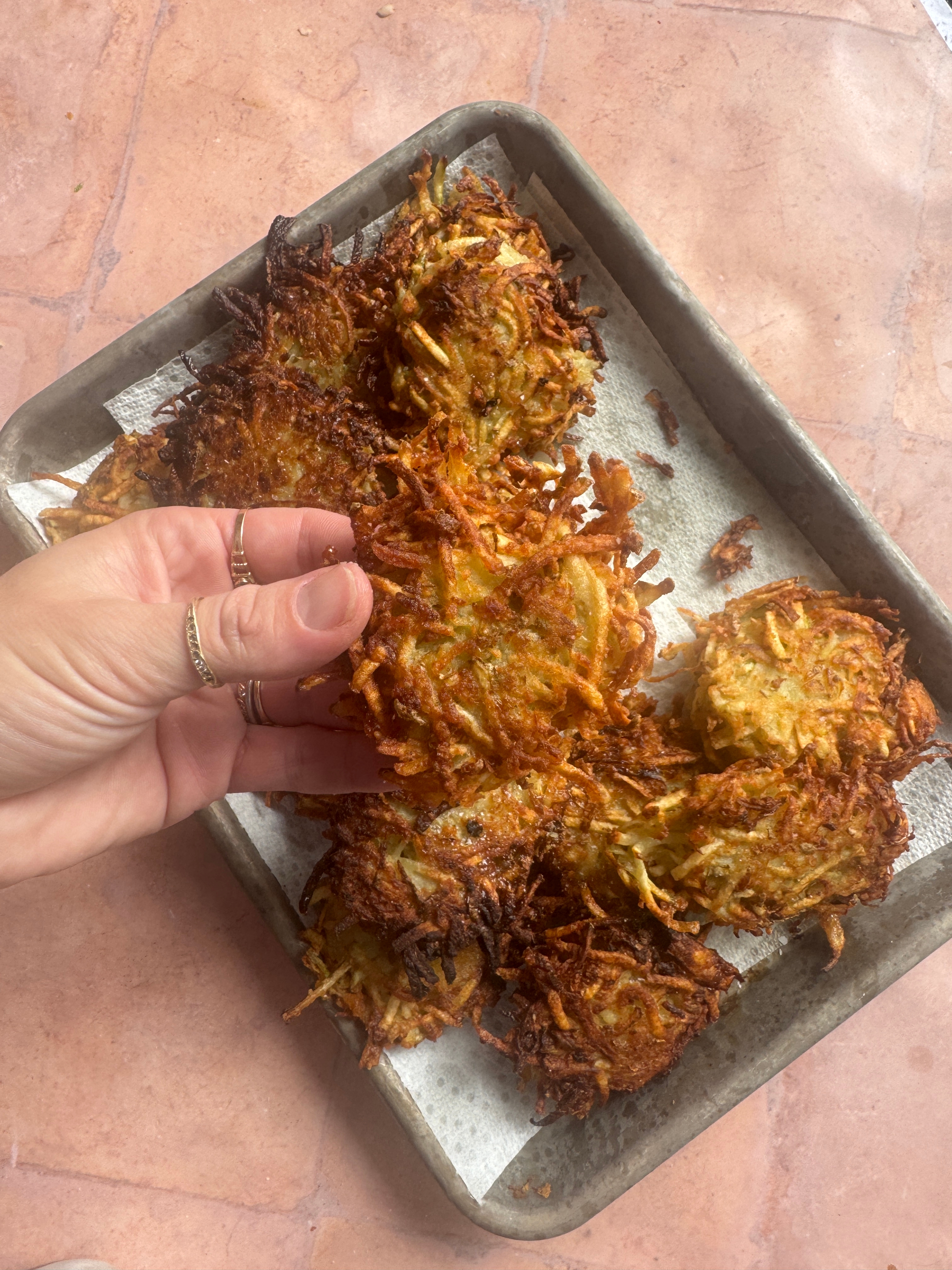 Person holding a crispy, homemade potato latke with a tray of more latkes on a paper-lined baking sheet