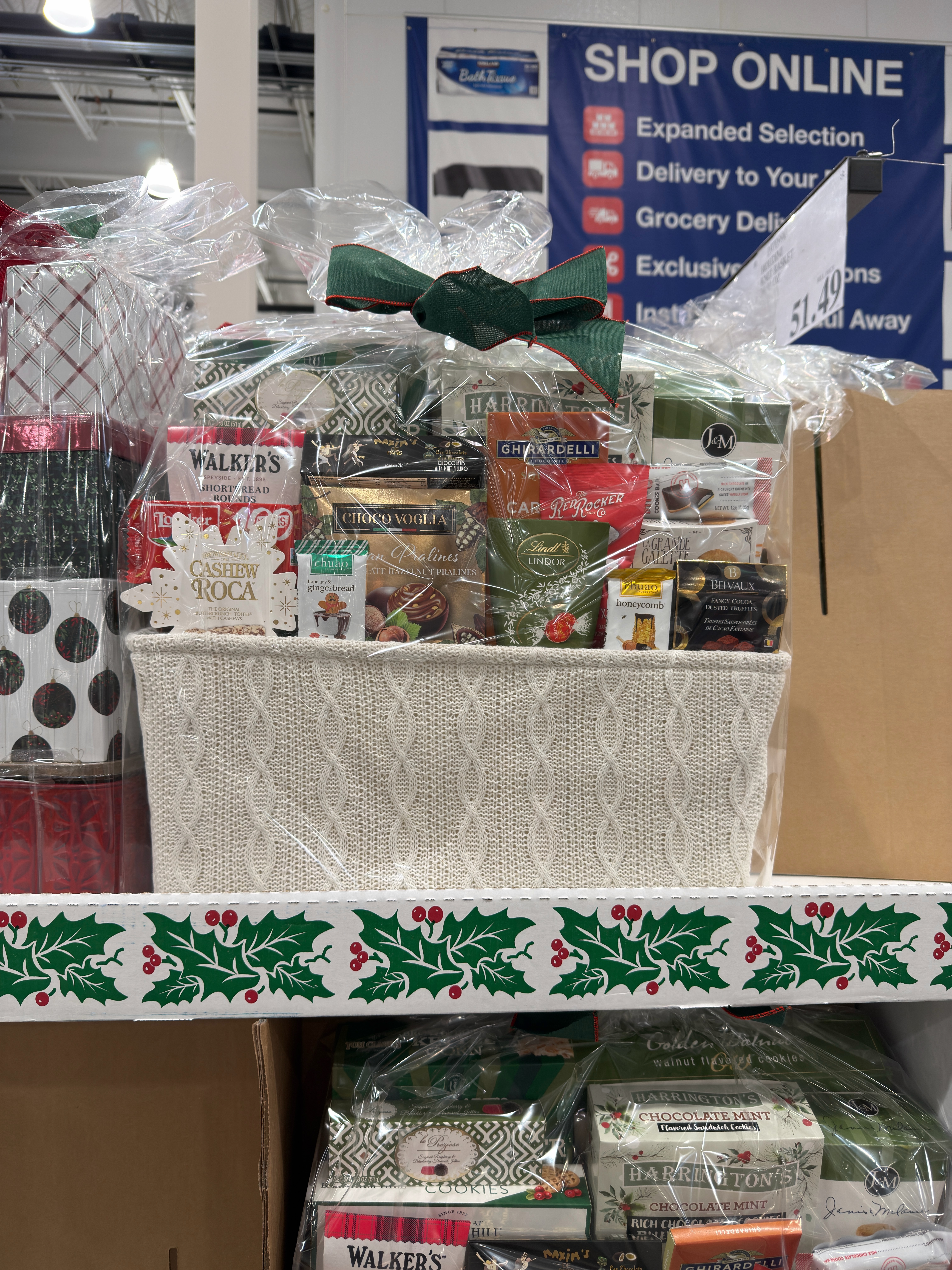 Gift basket filled with gourmet snacks like cookies and cocoa. It's wrapped in clear plastic with a green bow, displayed on a holiday-themed shelf