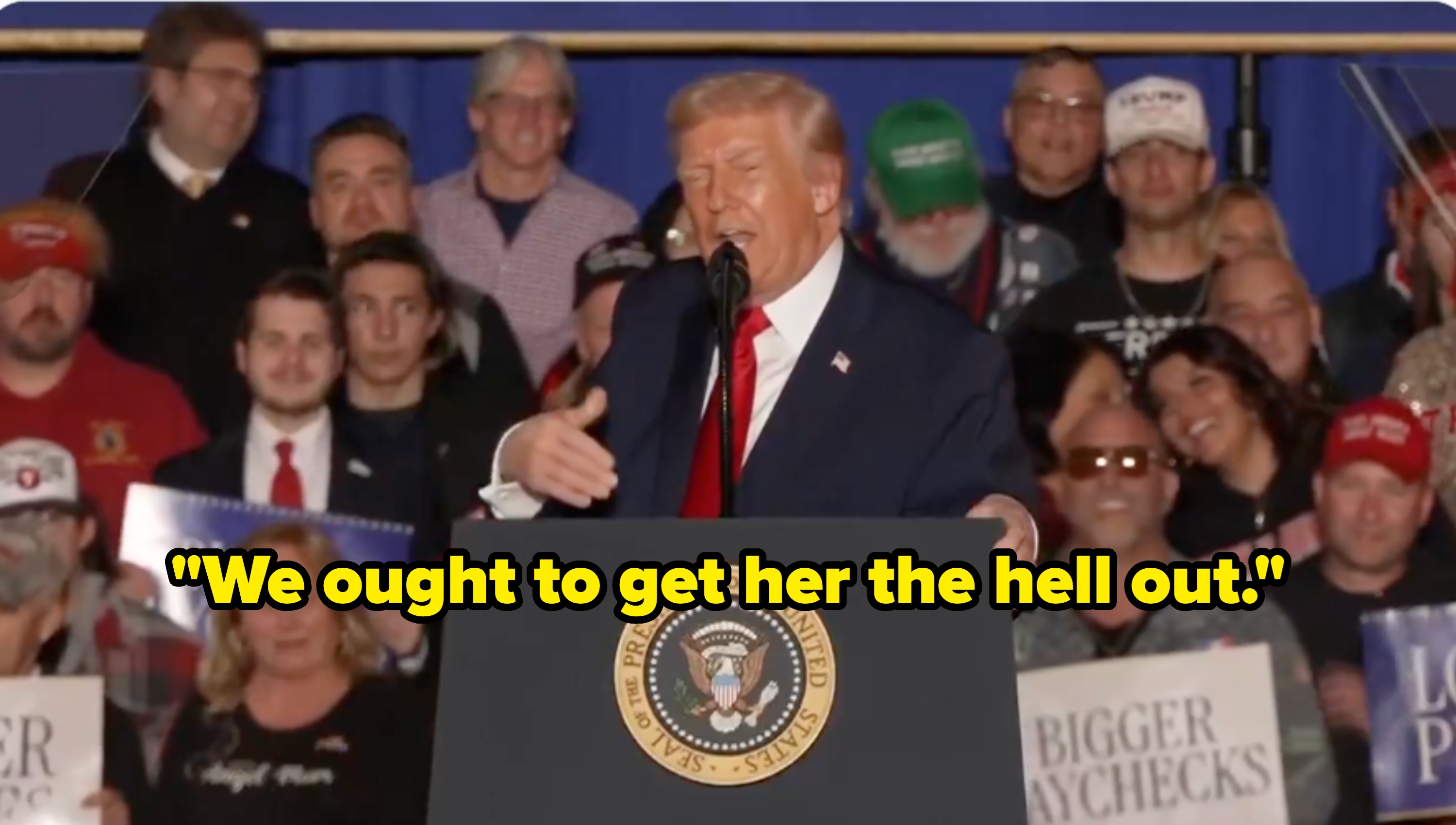 Offentlig person taler ved et podium med presidentseglet, omgitt av en folkemengde som holder skilt