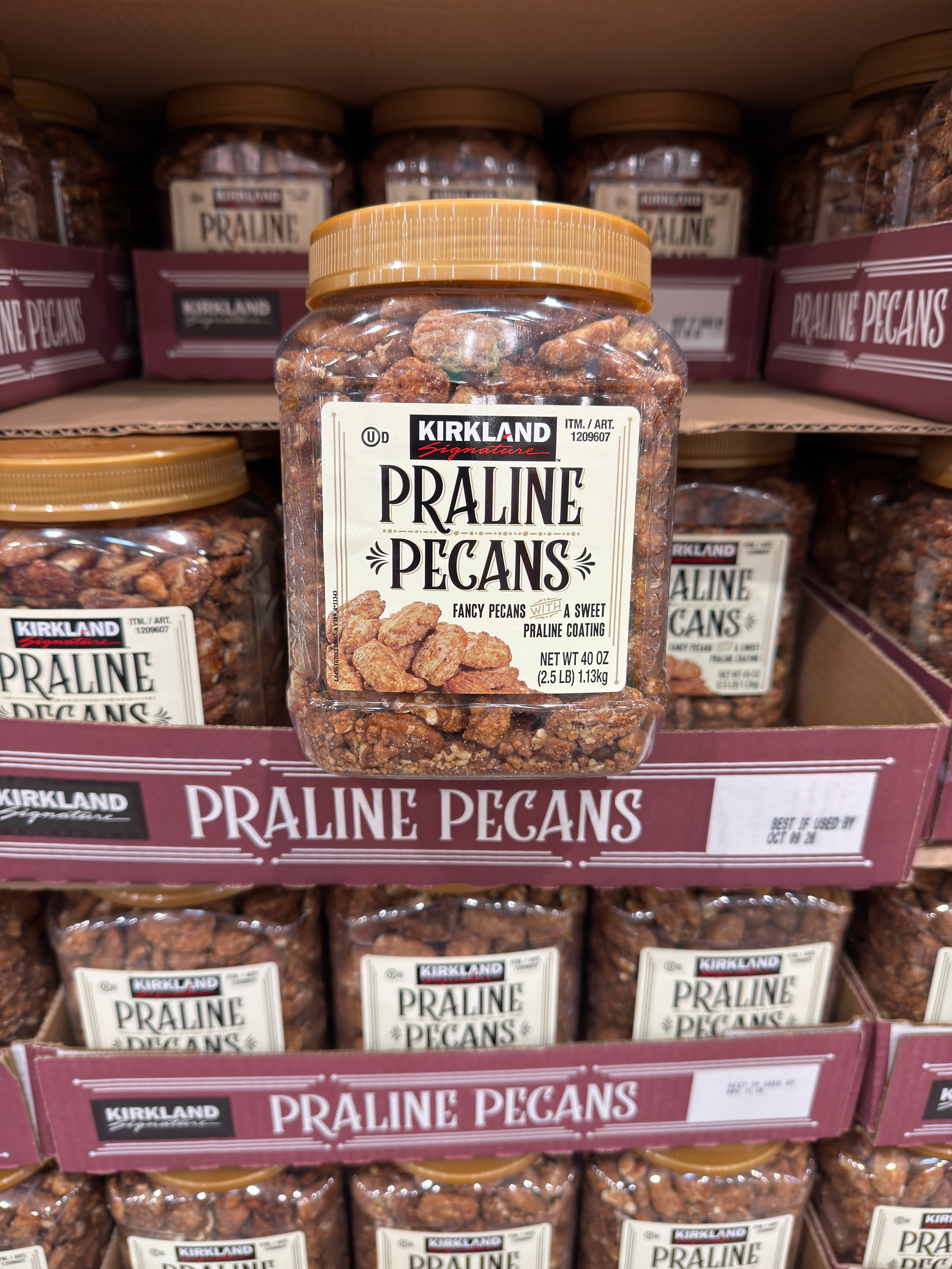 Jar of Kirkland praline pecans on a store shelf, surrounded by similar jars stacked with visible labels