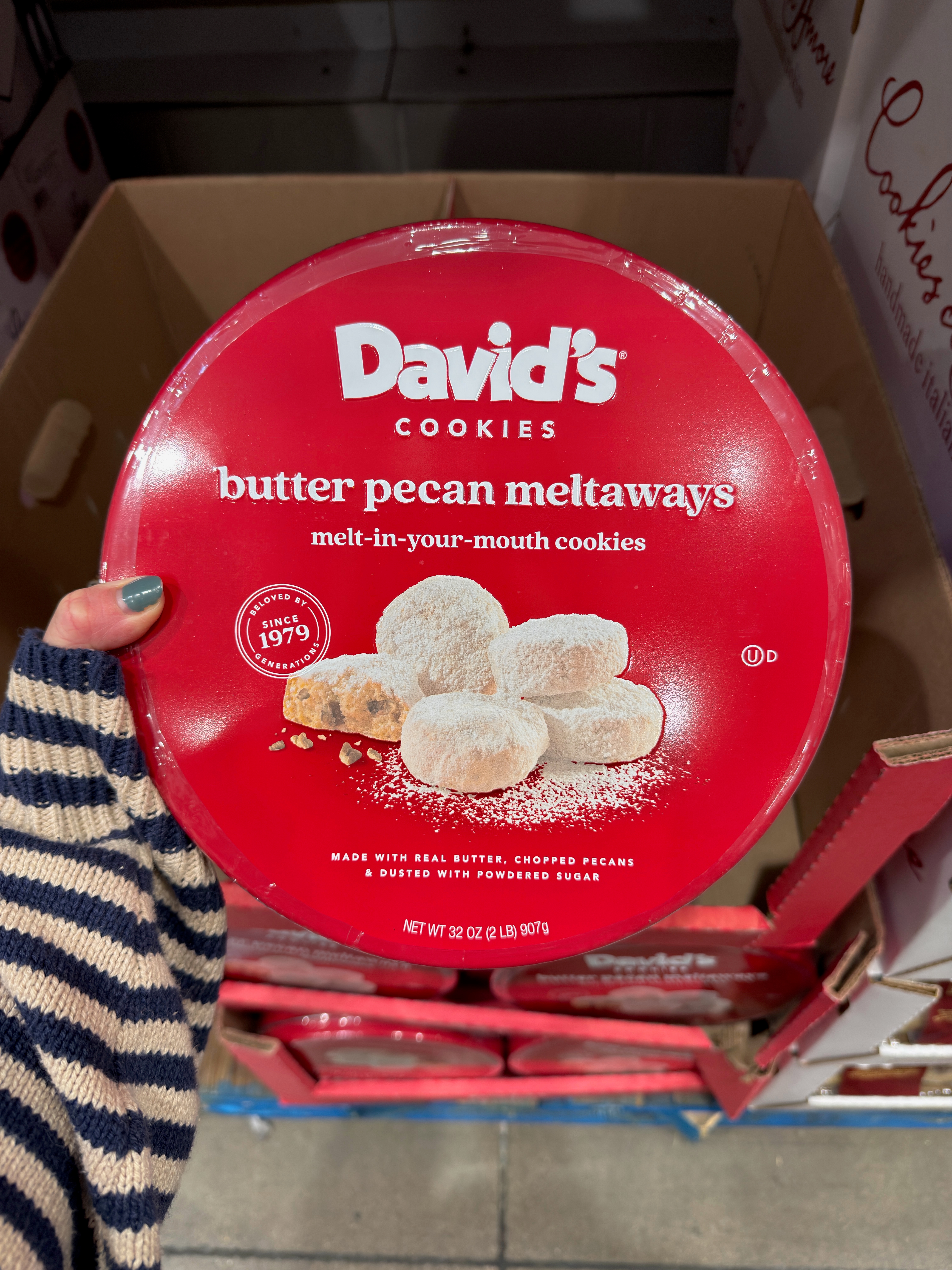 Round box of David's Cookies butter pecan meltaways, shown held over store shelf, featuring text and image of powdered cookies