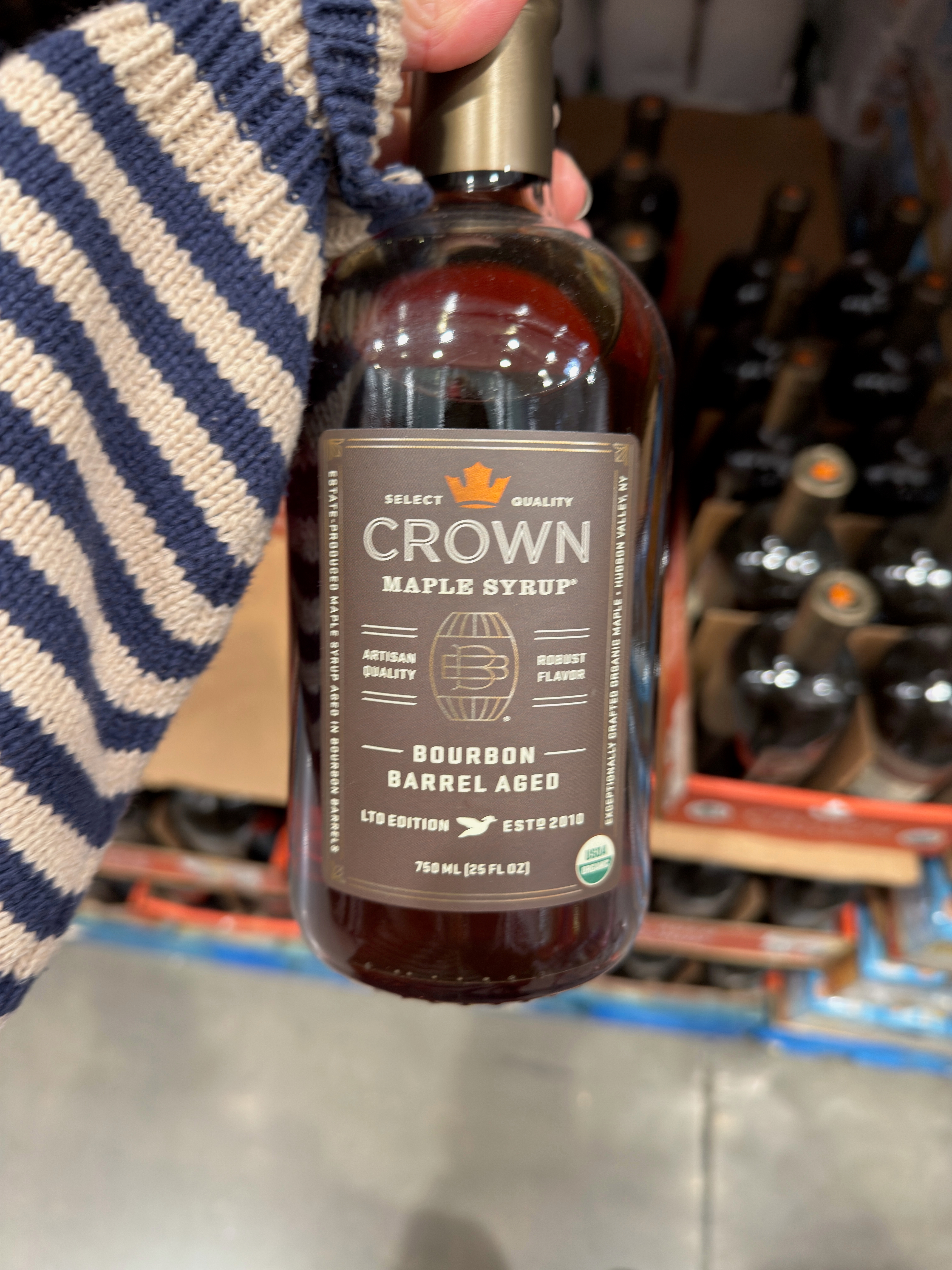 A hand holds a bottle of Crown Maple Syrup labeled as bourbon barrel aged, surrounded by other bottles on a store shelf