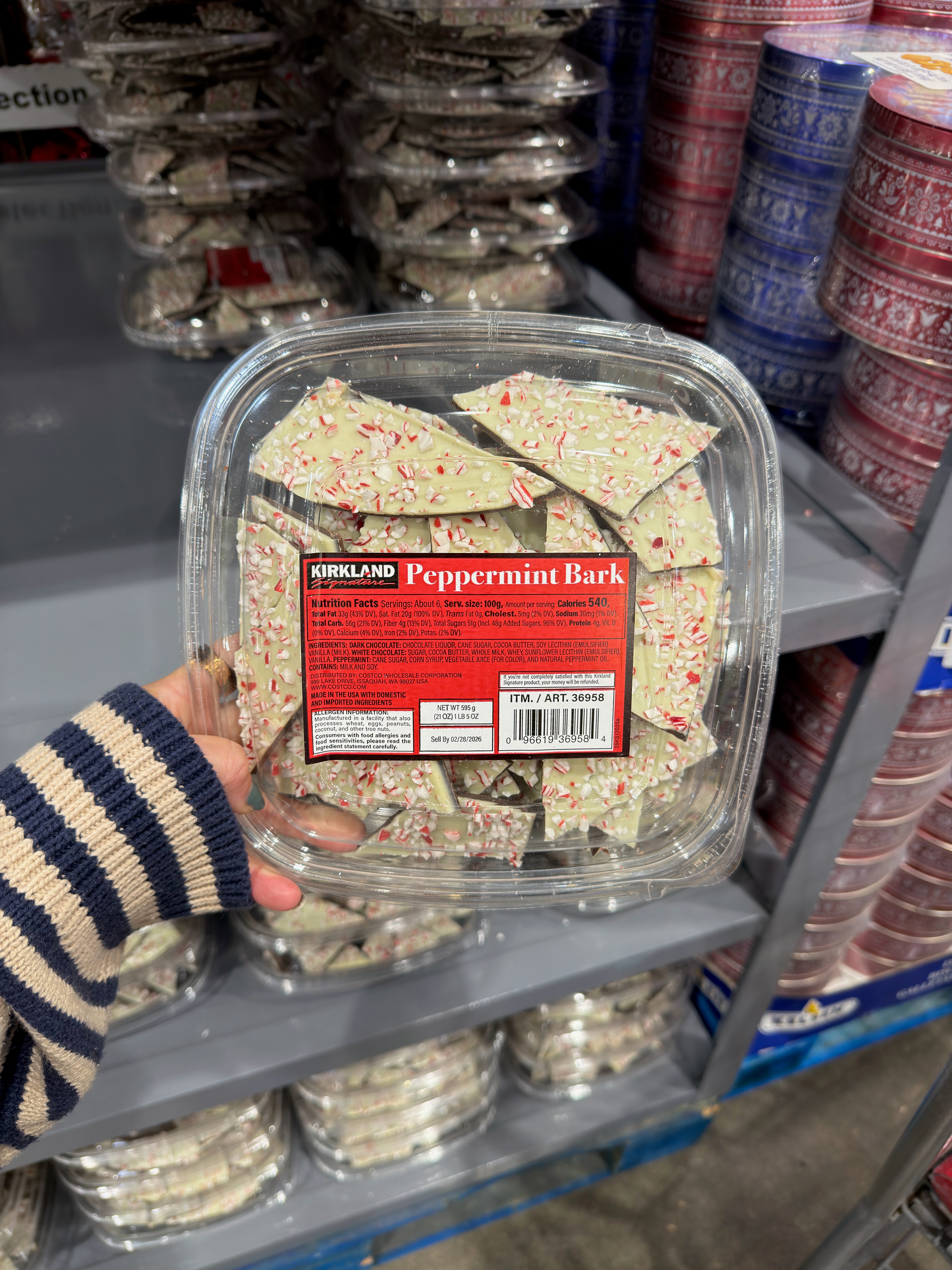 Person holding a container of Kirkland peppermint bark in a store aisle with stacked festive tins in the background