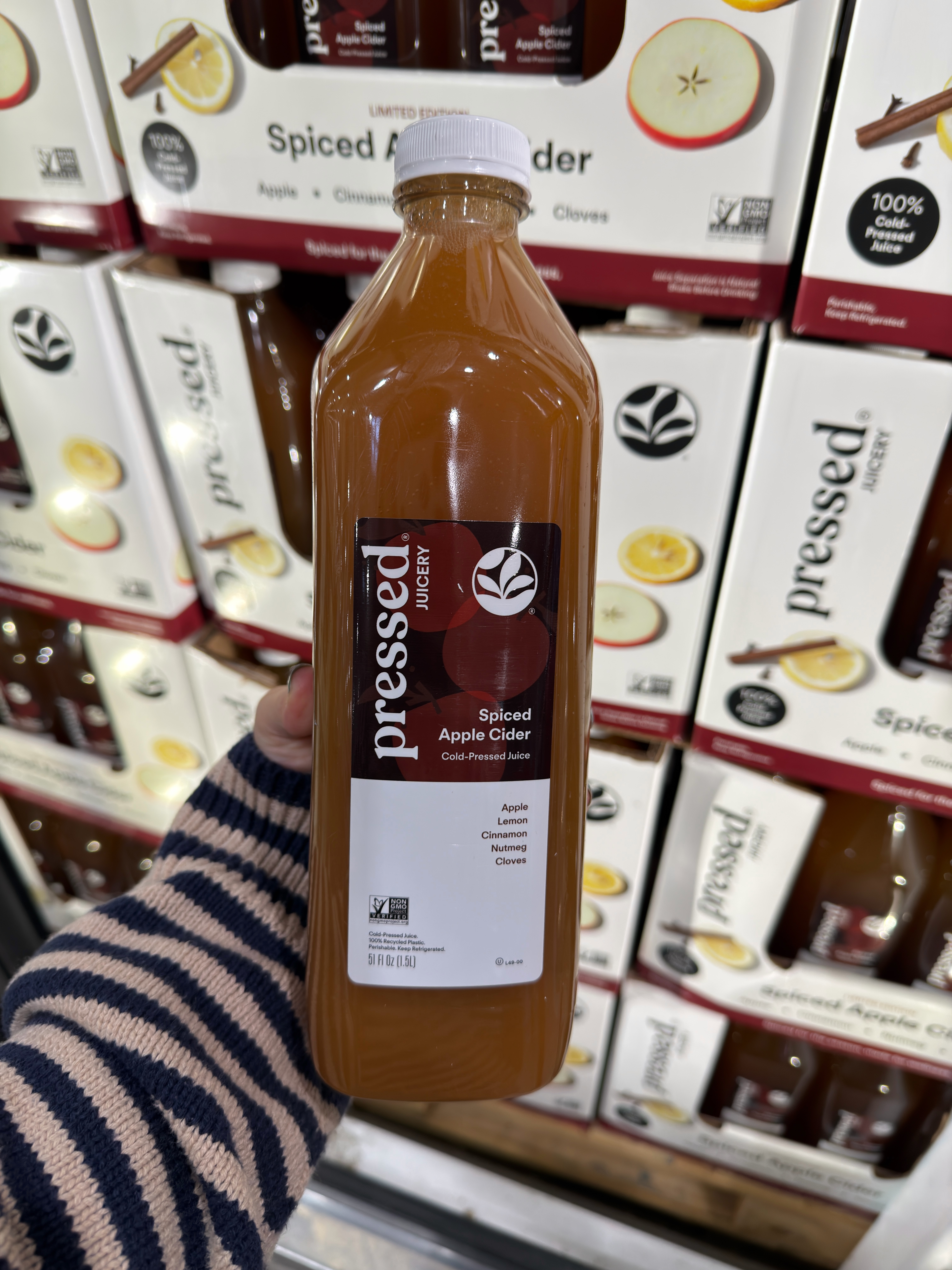 Person holding a bottle of pressed spiced apple cider in a store aisle with more bottles on shelves in the background