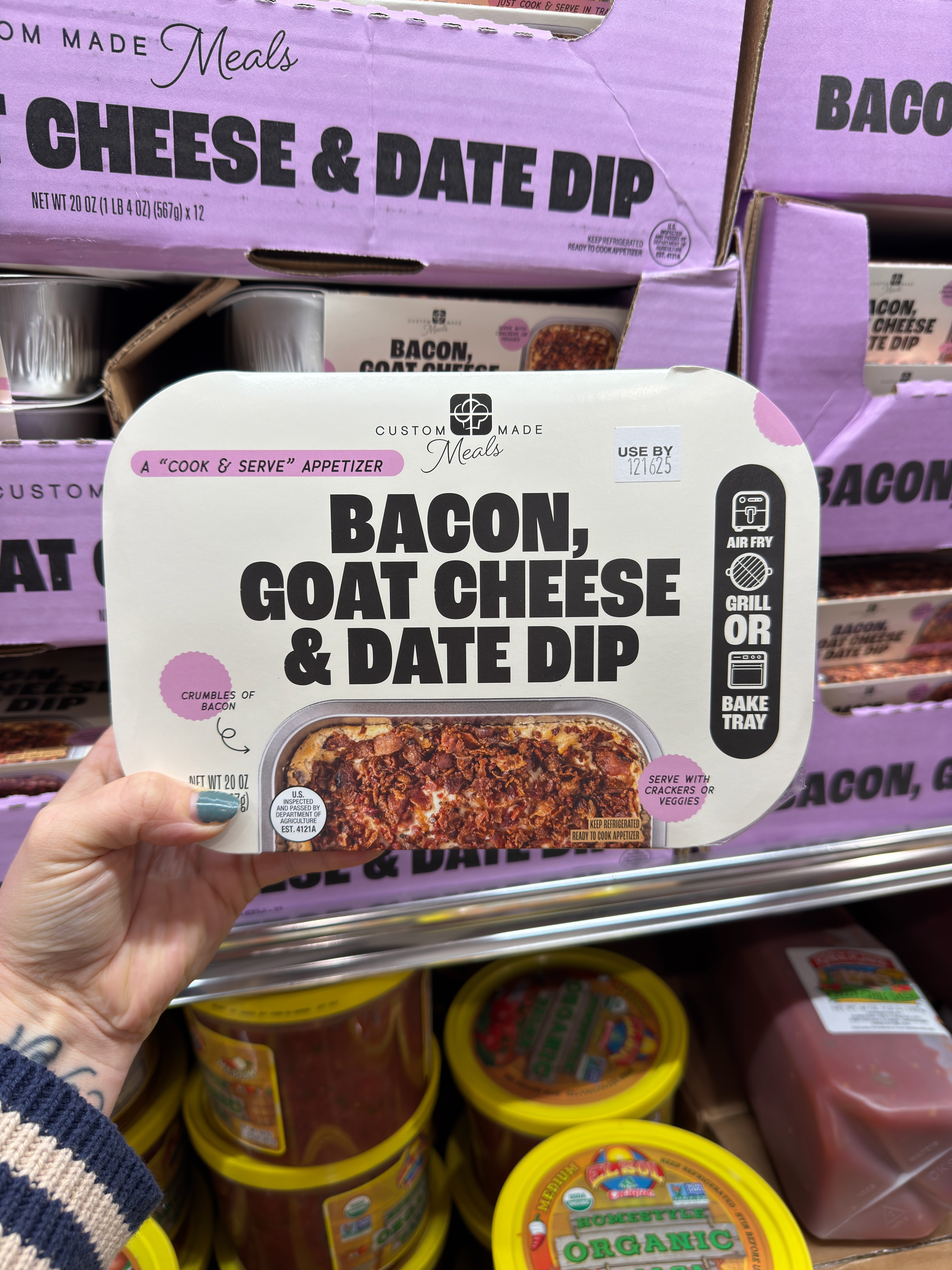 Person holding a packaged Bacon, Goat Cheese &amp;amp; Date Dip in a store. Background shows more dip containers on the shelf