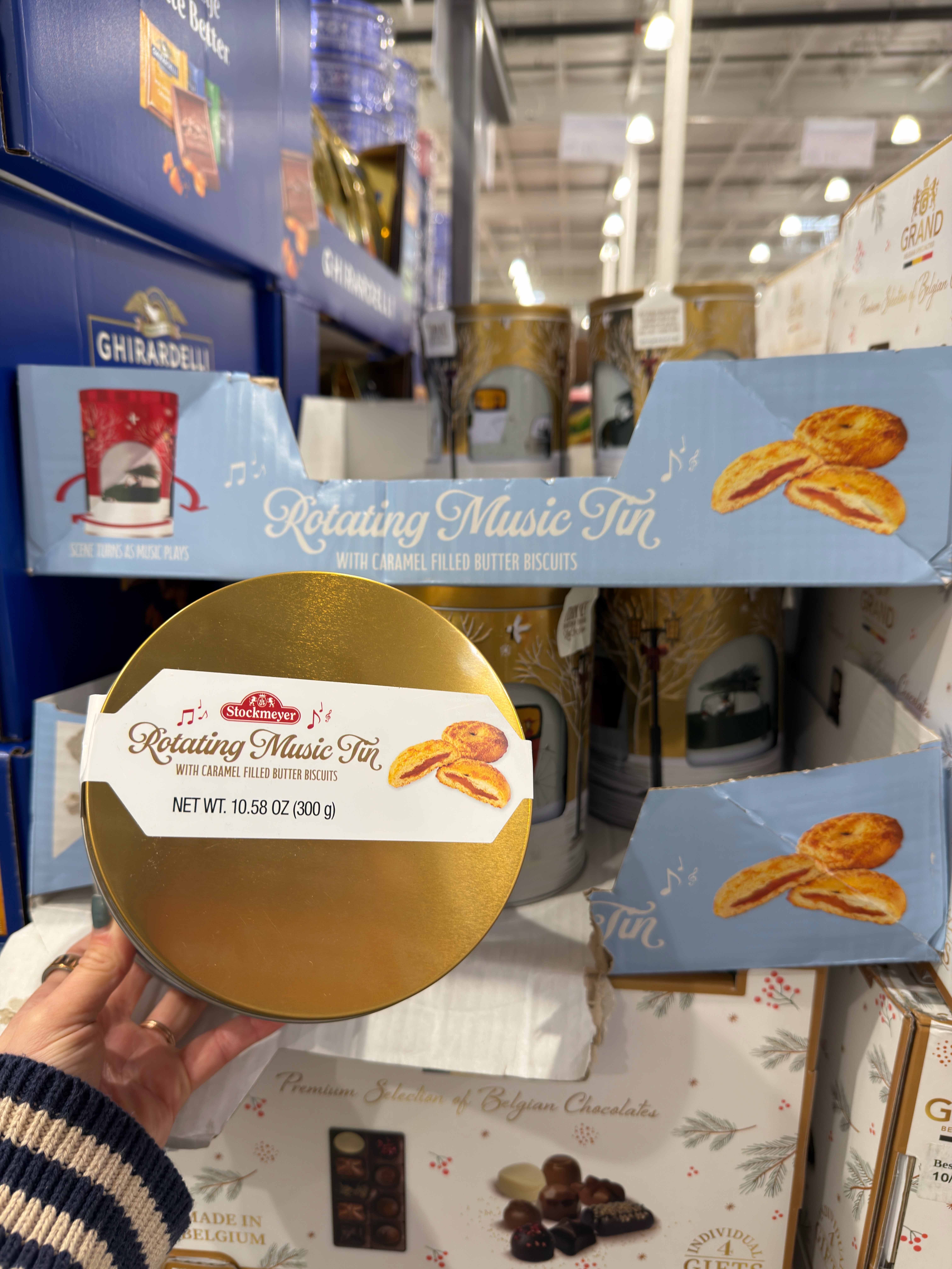 A person holds a tin of cookies labeled "Rotating Music Tin" in a store aisle, with packaging featuring caramel-filled butter biscuits