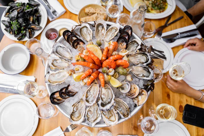 A table with a seafood platter featuring oysters, shrimp, and mussels on ice, surrounded by dishes, cutlery, and people holding wine glasses