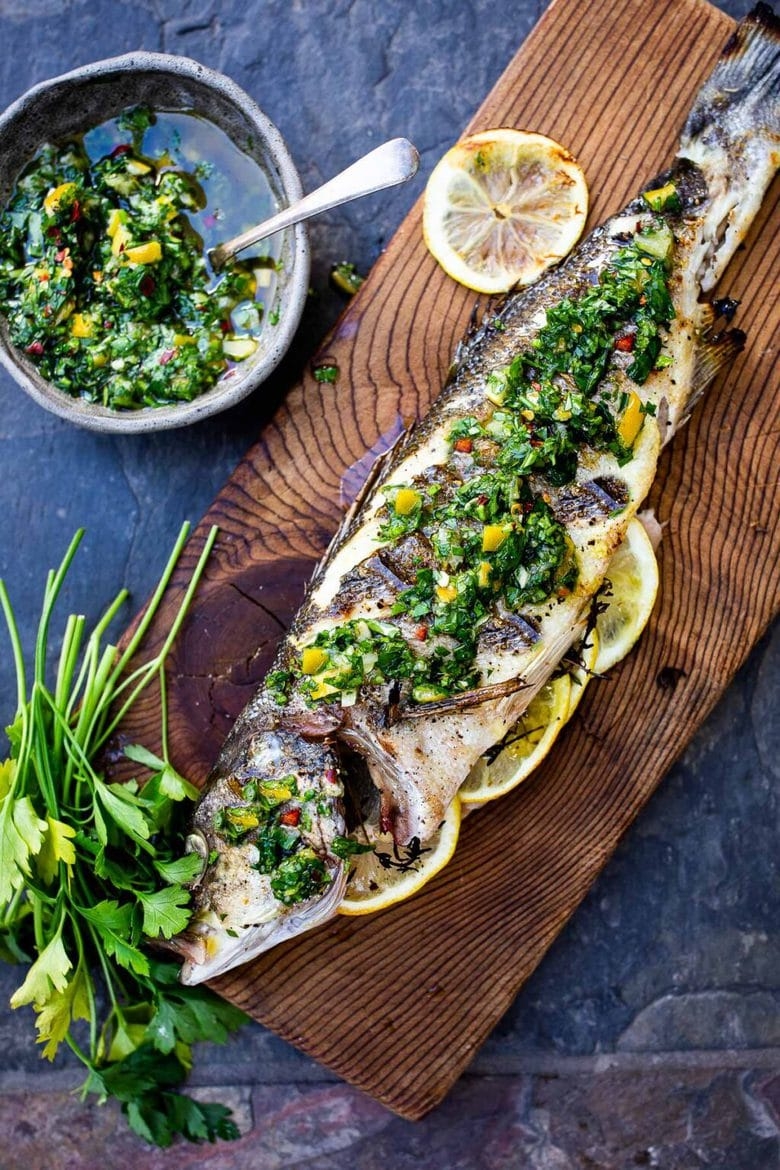 Grilled fish topped with herbs and lemon slices on a wooden board, served with herb sauce and fresh parsley on the side
