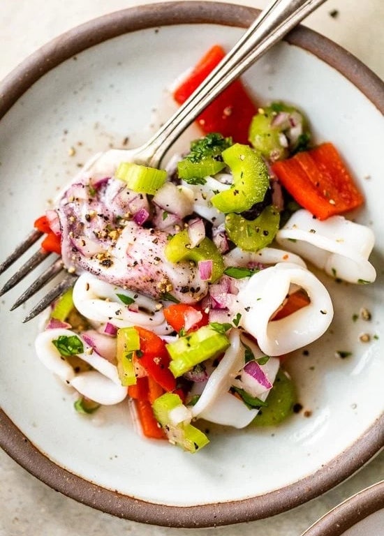 A fork rests on a plate of fresh calamari salad with diced vegetables, including celery and red bell peppers, garnished with herbs