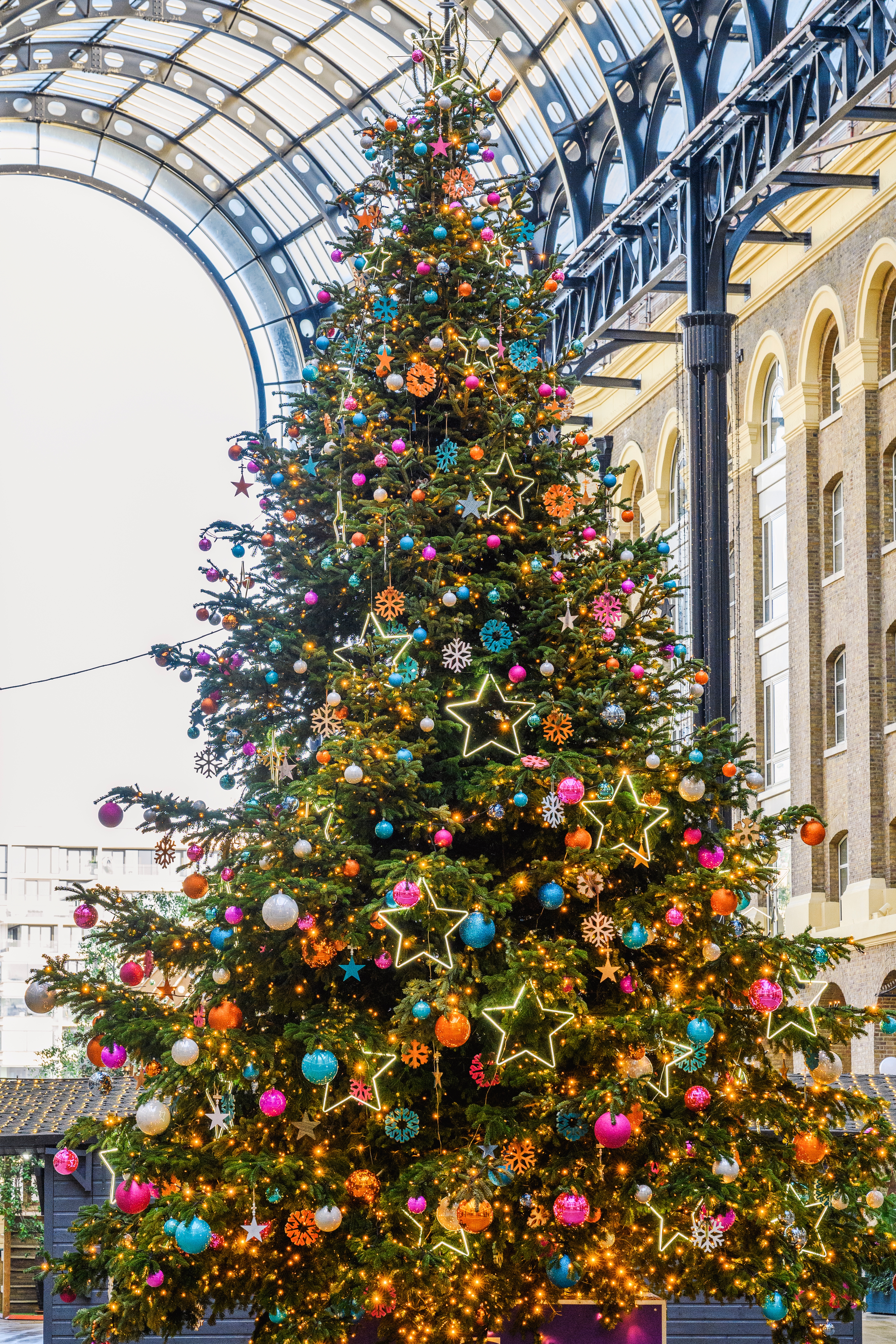 Stort pyntet juletre med stjerner, pyntegjenstander og lys står innendørs under et buet glasstak