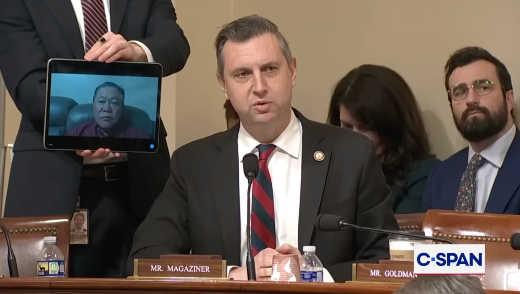 En person snakker under en høring mens en annen holder et nettbrett som viser en virtuell deltaker. C-SPAN logo synlig