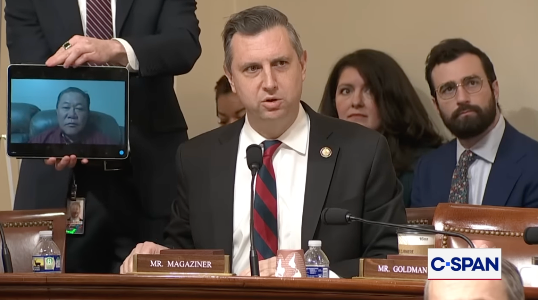 En kongresshøring med tjenestemenn; man holder et nettbrett som viser en person på en videosamtale