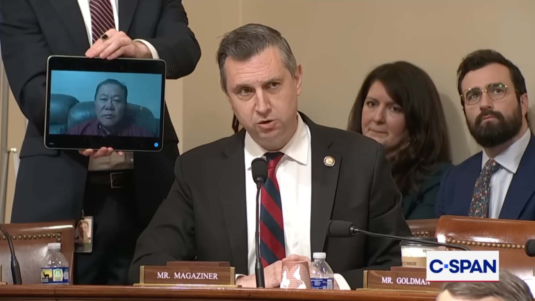 En mann i dress snakker under en formell høring, med et nettbrett som viser en annen person på en videosamtale som holdes av noen ved siden av seg. C-SPAN logo synlig