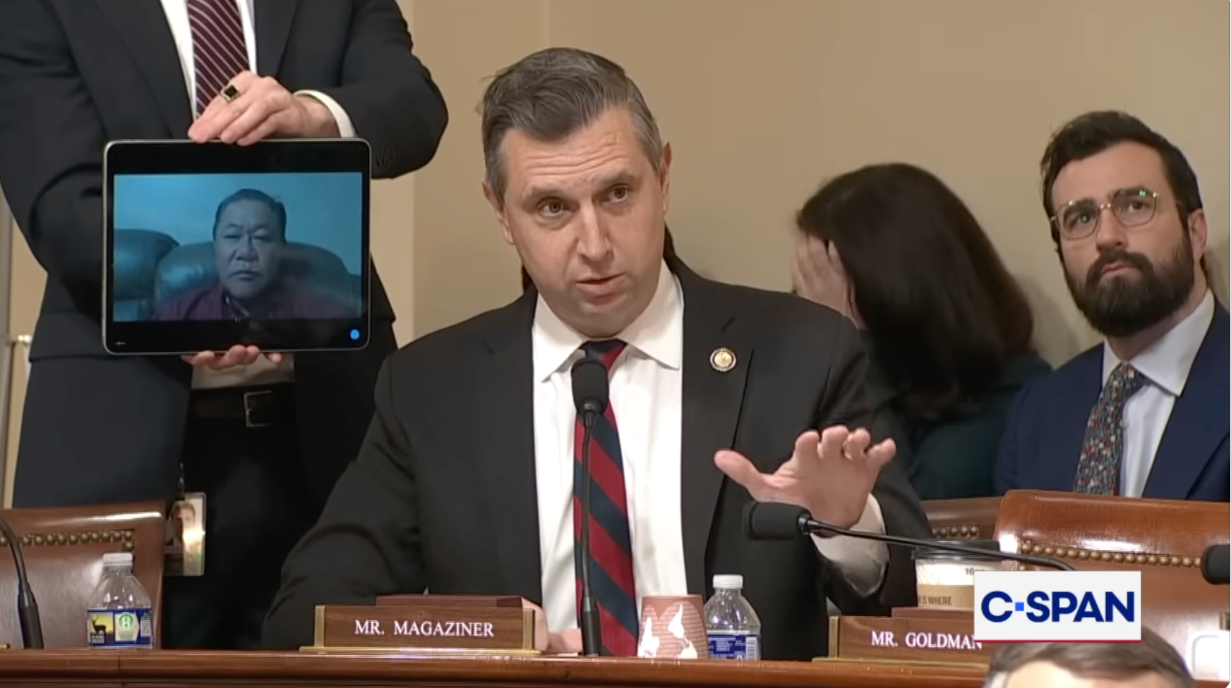 En mann i dress taler under en kongresshøring, med en person på skjermen ved siden av seg. C-SPAN logo synlig