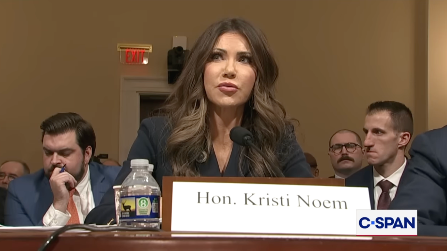 Hon. Kristi Noem taler under en kongresshøring, sittende ved et bord med navneskilt. Flere personer sitter bak henne. C-SPAN-logoen er synlig