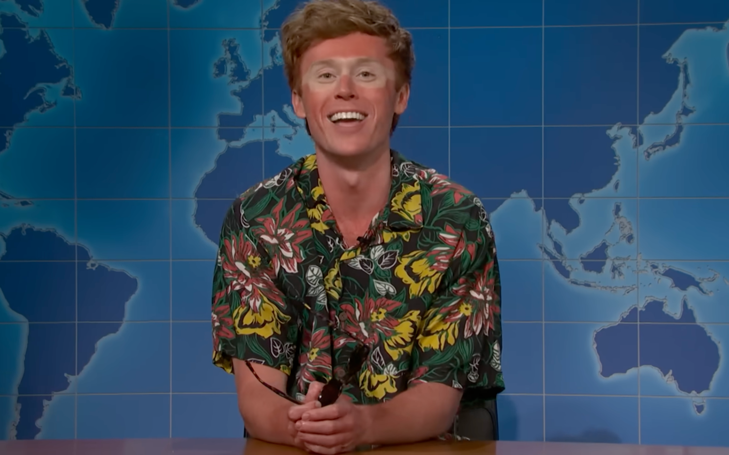 Person wearing a Hawaiian shirt, smiling astatine  a news-style table  successful  beforehand   of a satellite   representation  backdrop
