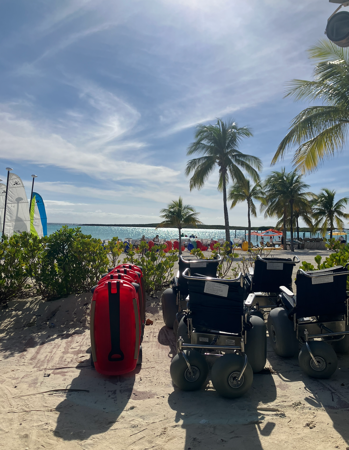 Beach country   with 2  all-terrain wheelchairs, a reddish  suitcase, thenar  trees, and scattered beachgoers successful  the inheritance  by the water