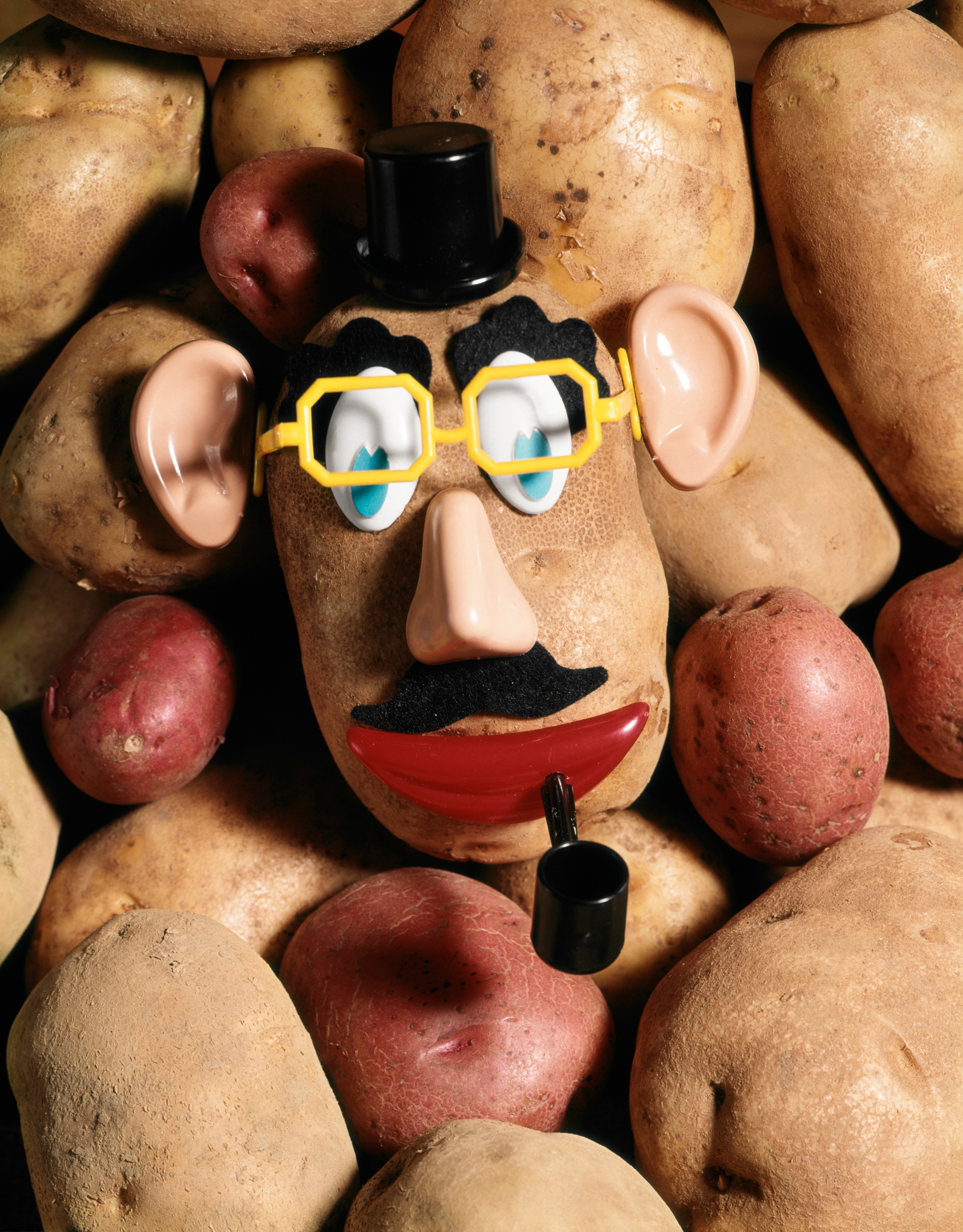 A Mr. Potato Head toy lies among real potatoes, featuring glasses, a mustache, a pipe, and a top hat, highlighting a playful mix of toy and reality