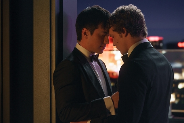 Two people in tuxedos share an intimate moment, leaning in with city lights in the background
