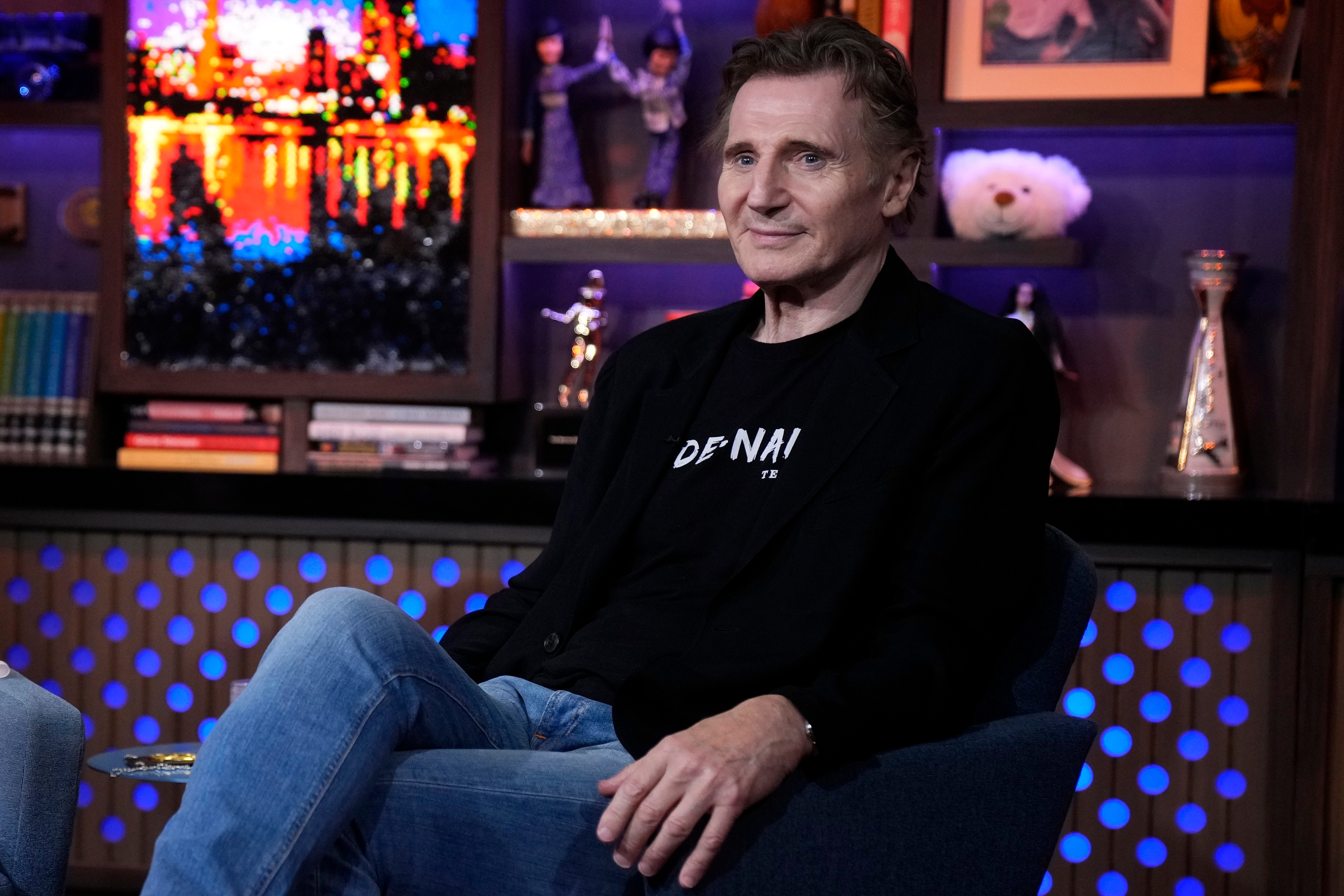 Liam Neeson sitter i et TV-studio, iført en uformell blazer og jeans, med en avslappet oppførsel