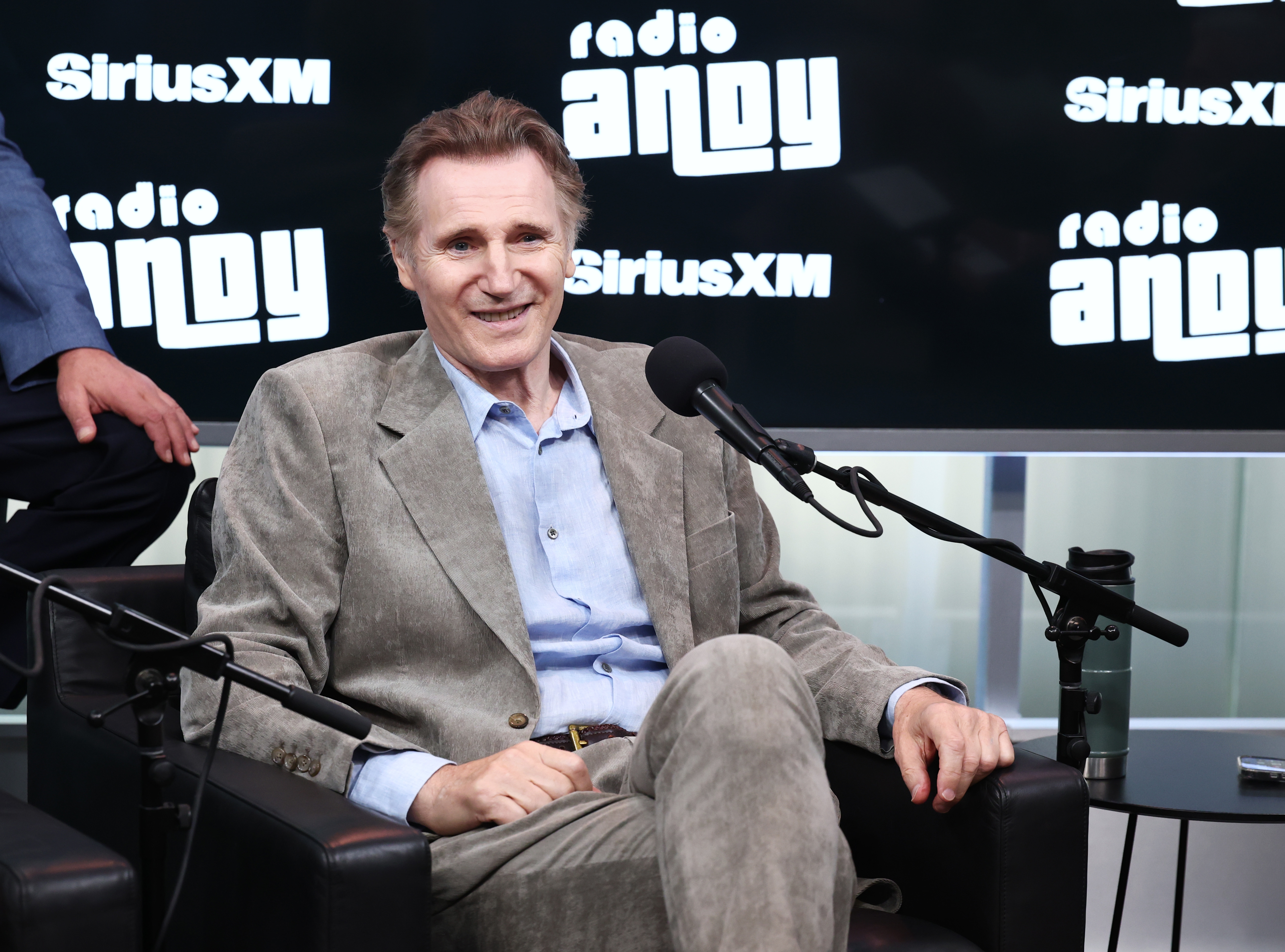Liam Neeson i dress og skjorte sitter smilende i et radiostudio med SiriusXM og Radio Andy-logoer i bakgrunnen