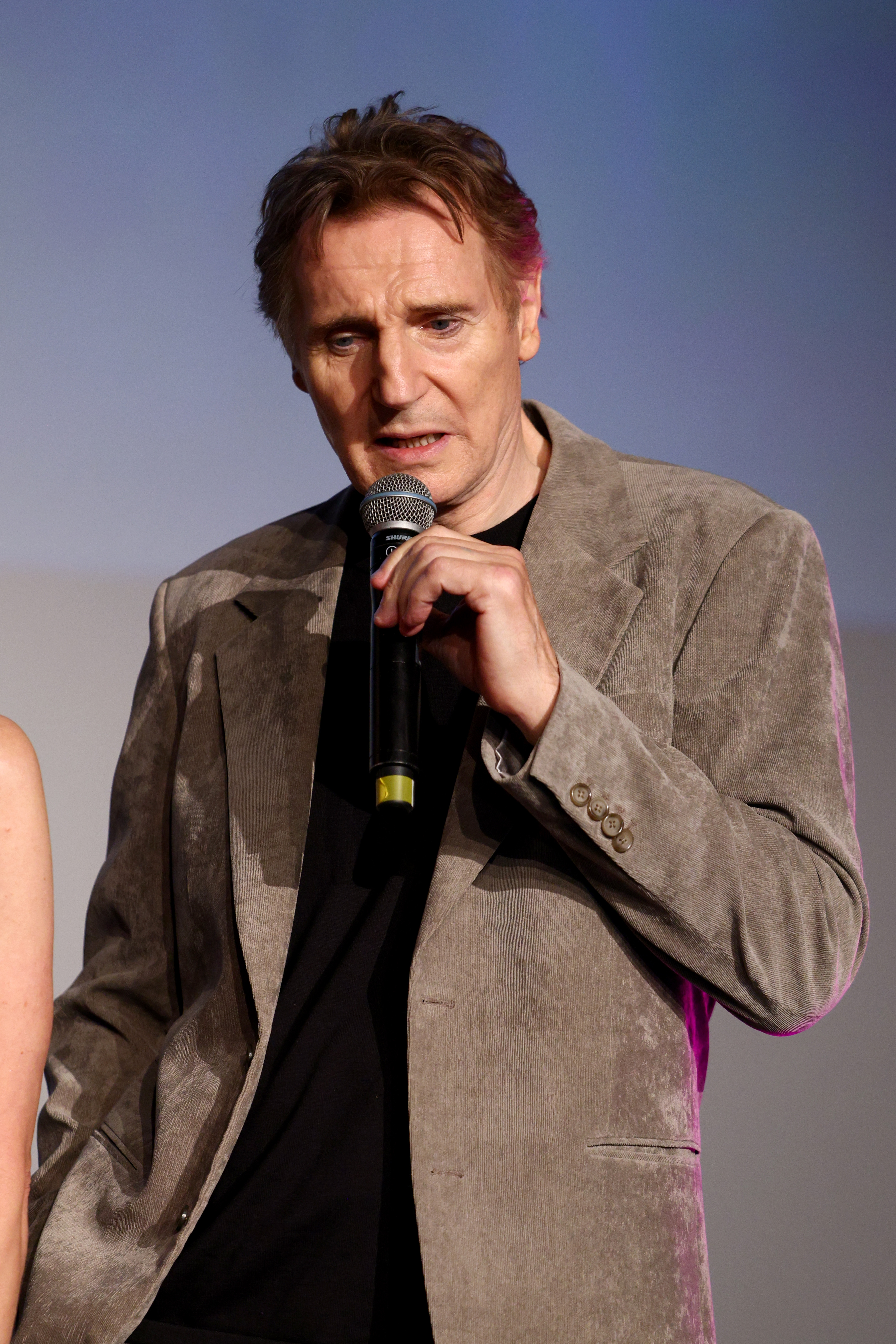 Liam Neeson i fløyelsjakke snakker inn i en mikrofon på scenen